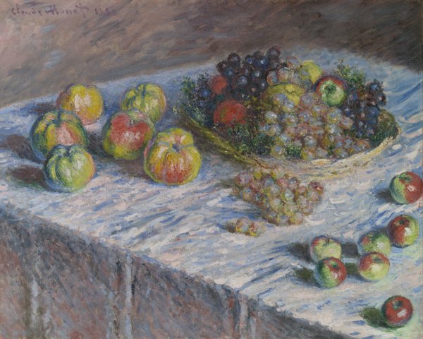 Apples and grapes rest in a shallow wicker basket on a table covered with a light-colored cloth. Additional fruit is scattered on the table.