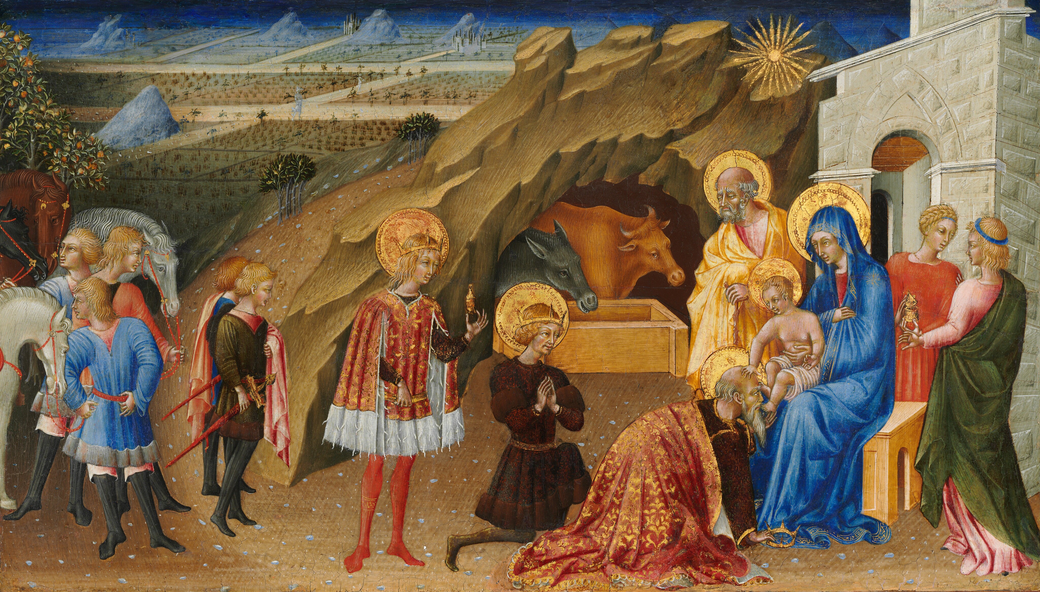 Three men with crowns and wearing gold-trimmed robes stand and kneel before a woman sitting on a wooden, box-like seat holding a baby to our right, in front of a rocky cave and landscape, in this horizontal painting. A balding man with a gray beard and hair, Joseph, and two younger people with blond hair stand around the woman, Mary, to our right, while a group of five young men or boys and three horses gather near the left edge. All the people have pale skin. Mary, the baby Jesus, Joseph, and the three men closest to them all have shimmering gold halos. One balding man with a long white beard kneels and kisses the baby’s feet. He wears a gold and crimson-red brocade-like robe over a burgundy-red garment, and he holds a pointed gold crown in his right hand, which rests on the ground. Behind him and to our left, a younger man with curly blond hair and goatee, wearing a wine-red tunic belted with gold and a gold crown on his head, holds his hands in prayer as he kneels down on one knee. The third stands to our left, cleanshaven with long blond hair. He wears a fur-lined, thigh-length scarlet-red robe and a gold crown, and he holds up a gold vessel in one hand. To our right, Mary wears a lapis-blue robe that covers her head and falls to her feet. The baby is covered only by a white cloth across his hips, and he has short blond hair. He touches the kneeling man’s head with one hand. His body is pudgy but his face has more adult-like, delicate features. Joseph stands at the woman’s far shoulder, wearing a butter-yellow robe over a rose-pink garment. His hands are gathered on a walking stick in front of his chest and his brows are furrowed with deep lines. One of the two younger people behind Mary, to our right, holds a lidded gold vessel. The group of five attendants to our left gather around cream-white, gray, or chestnut-brown horses. These blond people wear garments of dark olive-green, coral-red, royal-blue, or rose-pink tunics over black stockings. The rocky, brown cave behind Mary and Jesus, to our right of center, is occupied by a donkey and an ox behind a wooden trough. The landscape beyond is segmented into quilt-like squares of stylized crops. The horizon line comes almost to the top edge of the panel beneath a narrow strip of vivid, cobalt-blue sky. A gold star like a sunburst with rays of light hovers over the woman and child.
