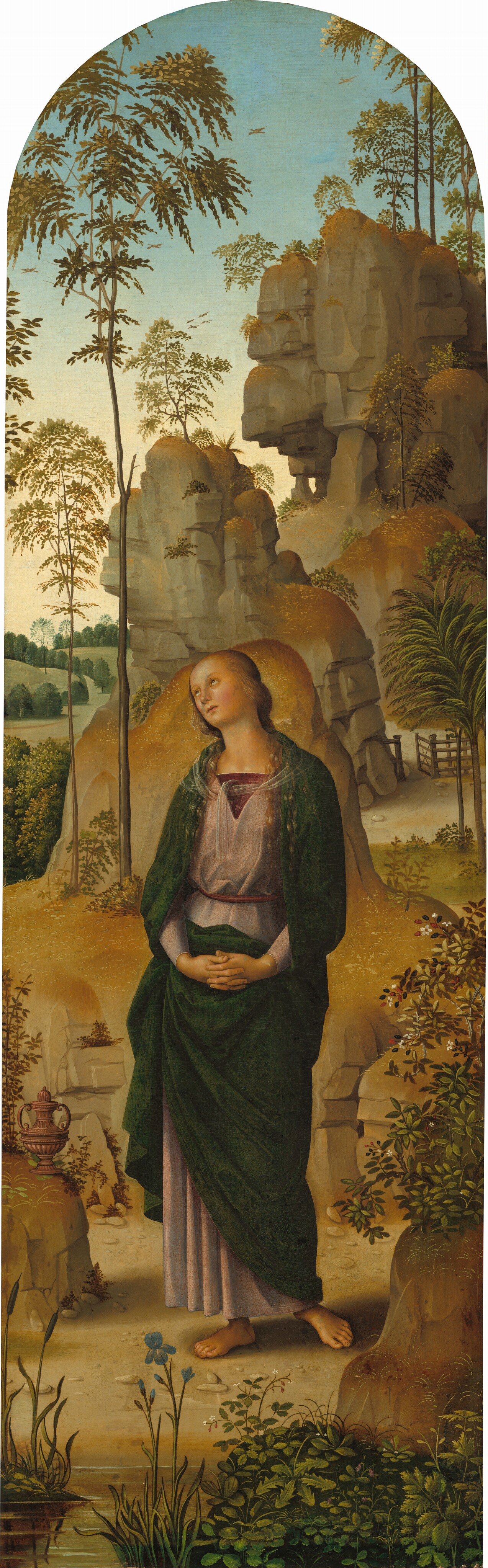 A young woman with pale skin and flushed cheeks stands in a landscape looking to our upper left in this round-topped vertical painting. Her body faces us in the center of the scene. Her fingers are interlaced at her waist, and her head tips slightly to our left. Honey-brown hair cascades over her shoulders, and her hazel eyes roll up under thin brows. She has a short nose and a small, pale pink mouth. A translucent scarf is tied around the full-length, pine-green mantle that drapes across her shoulders and wraps around the V-neck of her rose-red robe. A wine-red inset fills the neckline, and a matching belt is tied around her waist. She stands barefoot on the dirt ground, which is scattered with pebbles. Plants grow and bloom, including a blue iris, in shallow water along the bottom edge of the composition. A lidded pitcher with a narrow neck and scrolling handles sits on a flat rock next to her right knee. Taller, peanut-brown rock formations pile up beyond her under a powder-blue sky. Tawny-brown and sage-green trees, leafy plants, and shrubs sprout among the rocks. Beyond the woman is an open gate in a wooden fence built into one of the rock formations to our right, and sage-green hills dotted with moss-green trees roll out along the left side of the painting.