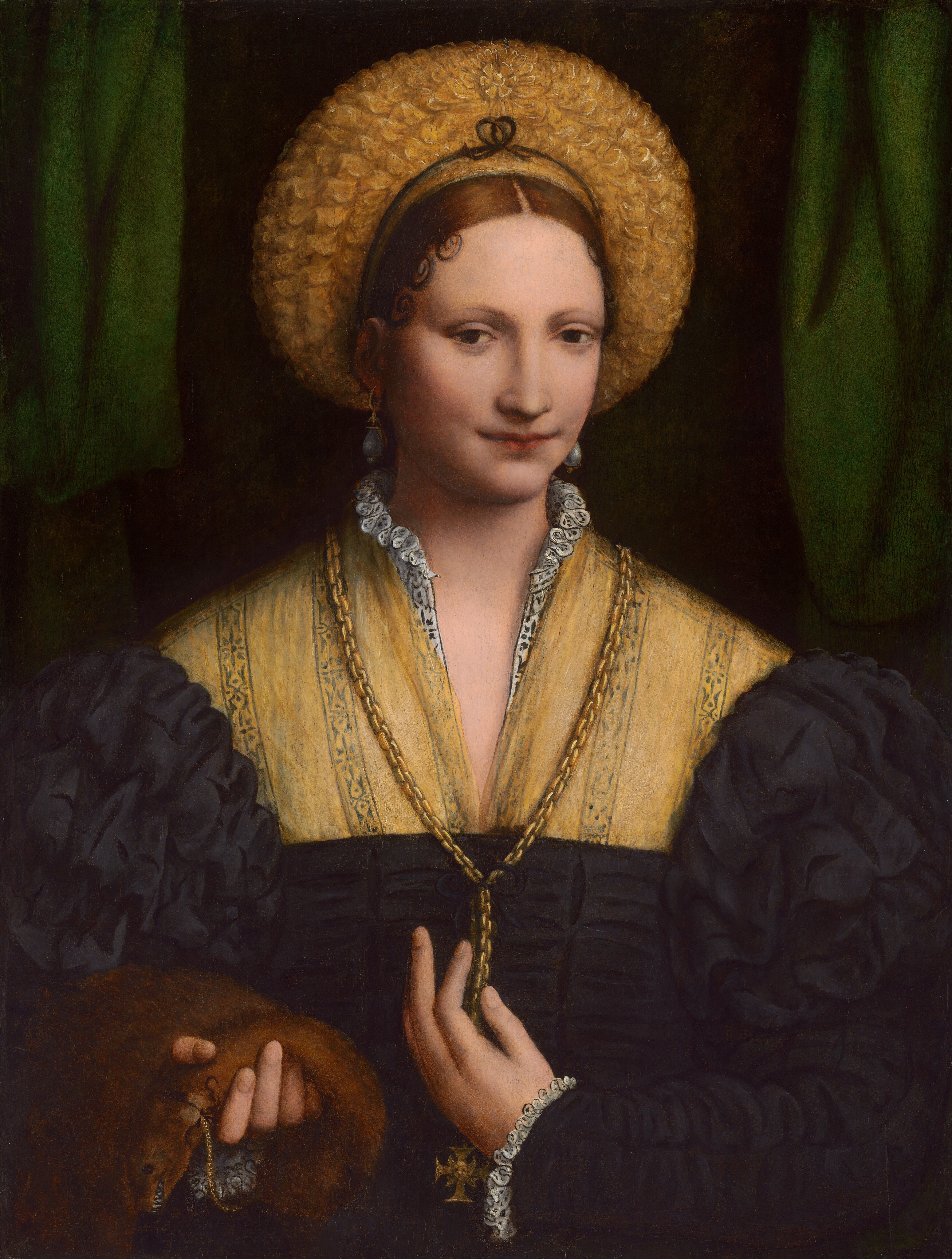 Shown from the waist up, a young woman with pale, peachy skin faces and looks directly at us in this vertical portrait painting. The subtly upturned corners of her mouth and the outer corners of her eyes are touched with shadows. Her delicate eyebrows are faintly painted over dark brown eyes. She has a long straight nose and an oval-shaped face framed by large teardrop pearl earrings. Her dark brown hair is parted down the middle and pulled back under a wide, gold-colored headdress, which creates a halo-like ring around her head. Precisely formed curls rest against her forehead over each temple. Her black dress has voluminous, puffy sleeves framing a sheer, butter-yellow panel covering her chest. White ruffles emerge at her neck and cuffs. A gold Maltese cross, which flares at the end of each arm, hangs from a thick gold chain that falls over her chest and down to her waist. She touches the chain with her left hand, on our right, and with the other she holds the body of a small furry brown animal with bared teeth. The background behind the woman is black down the center and a dark emerald-green curtain falls to either side.