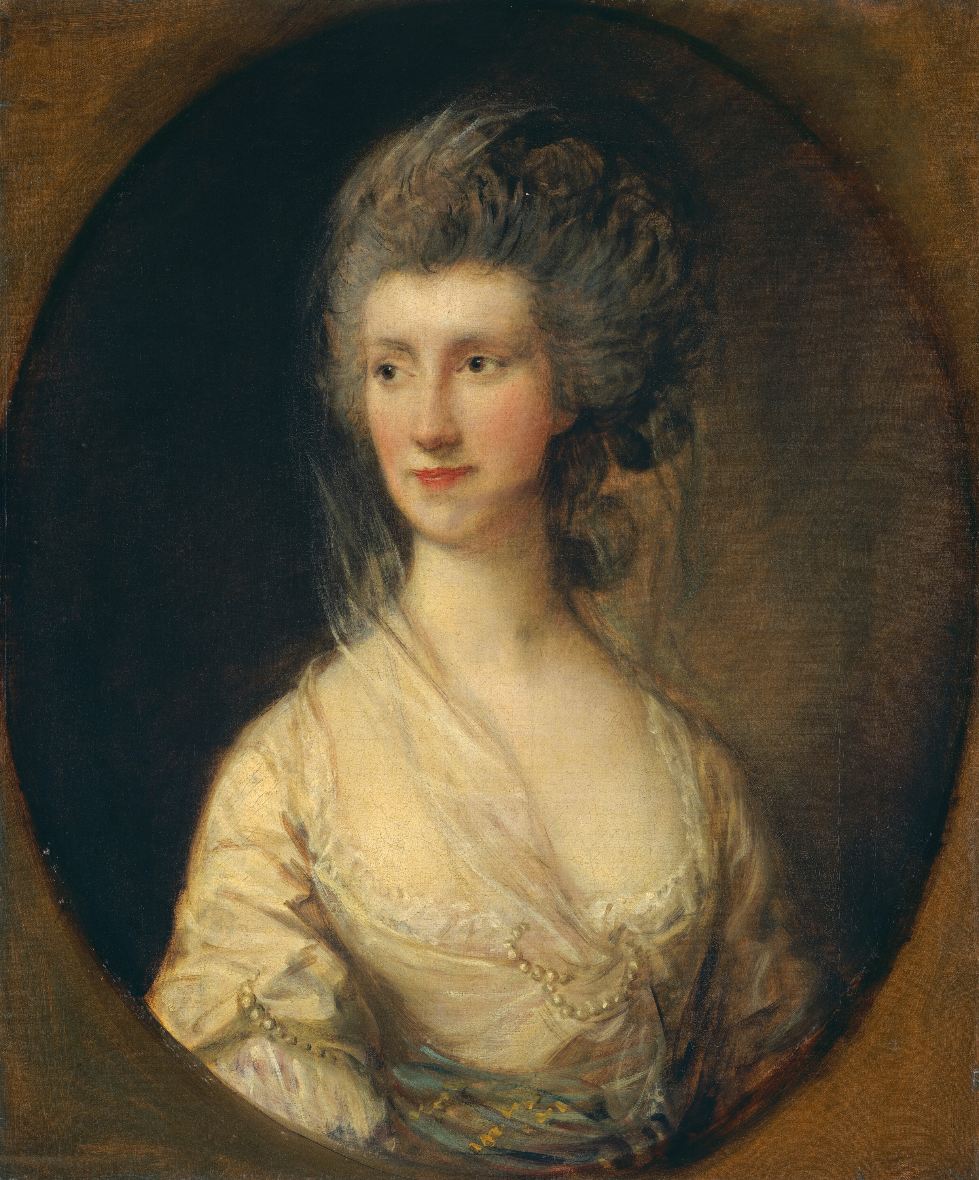 A pale-skinned woman is shown from the waist up within an oval opening in this vertical portrait painting. The woman’s body is angled to our right, but she turns her face to look off to our left with gray eyes. She has a thin, oval-shaped face, a long nose, and her dark pink lips are closed. Her ash-brown hair is swept up and back from her forehead, and some curls hang down the back of her neck. A sheer veil drapes over her hair and down around her sloping shoulders. The bodice of her ivory-yellow gown is cut low and lined with lace. Pearls ornament the front of the dress and part of the sleeve closer to us, and an aquamarine-blue cloth with gold touches is tied around her waist. The corners around the oval opening are painted to look like wood in shades of warm brown. The background behind the woman is nearly black to our left and it lightens to earthy brown to the right. The portrait is loosely painted, especially in the hair, clothing, and background.