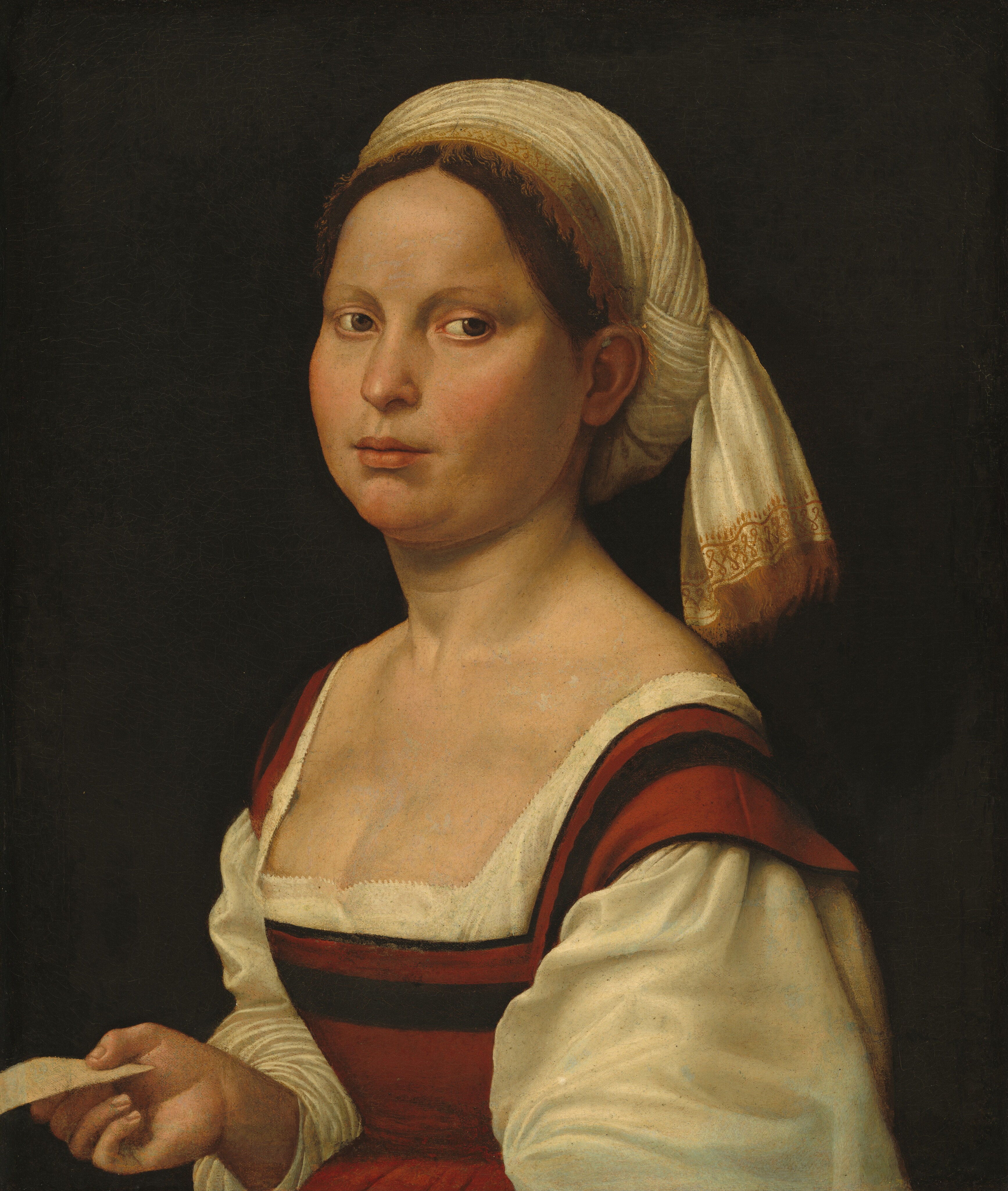 This painting shows the upper half of a woman's body, her torso turned to the left and her head turned slightly back to look towards the right. She has pale skin and brown hair that is parted in the middle and mostly covered by a white headscarf with a decorative golden edge and gold fringe. Her facial features include pale, arched eyebrows, rosy cheeks, brown eyes, and a small pink mouth in a neutral expression. Her clothing consists of a red dress with a low square neckline and a black stripe, with long white sleeves and a white undershirt poking through around the neckline. The woman holds a small piece of paper in her right hand. The background is solid black.