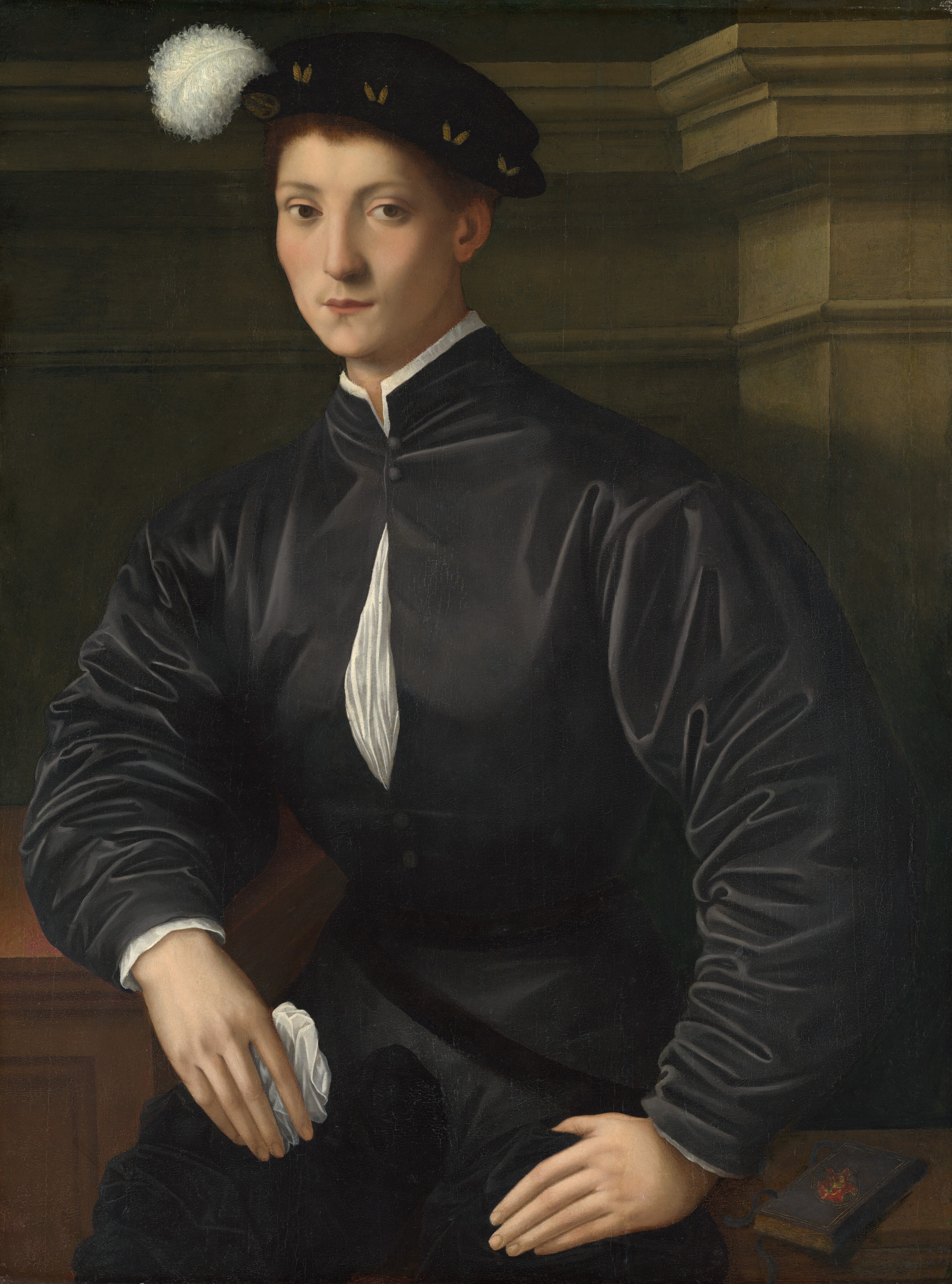 The painting shows a person sitting with their body slightly turned to the left and their head turned towards us. They have a fair complexion, with high cheekbones, a long nose, and thin pink lips in a neutral expression. They have short, light brown hair mostly covered by a dark hat with a white plume. The person wears a dark garment with puffed sleeves, a white collar, and a white strip on the chest. They rest their left hand on their knee while their right arm rests on a wooden surface next to them, their right hand holding a white cloth. In the bottom right corner, a small black book sits on a low wooden surface. In the background is a dark gray wall with decorative molding and paneling.