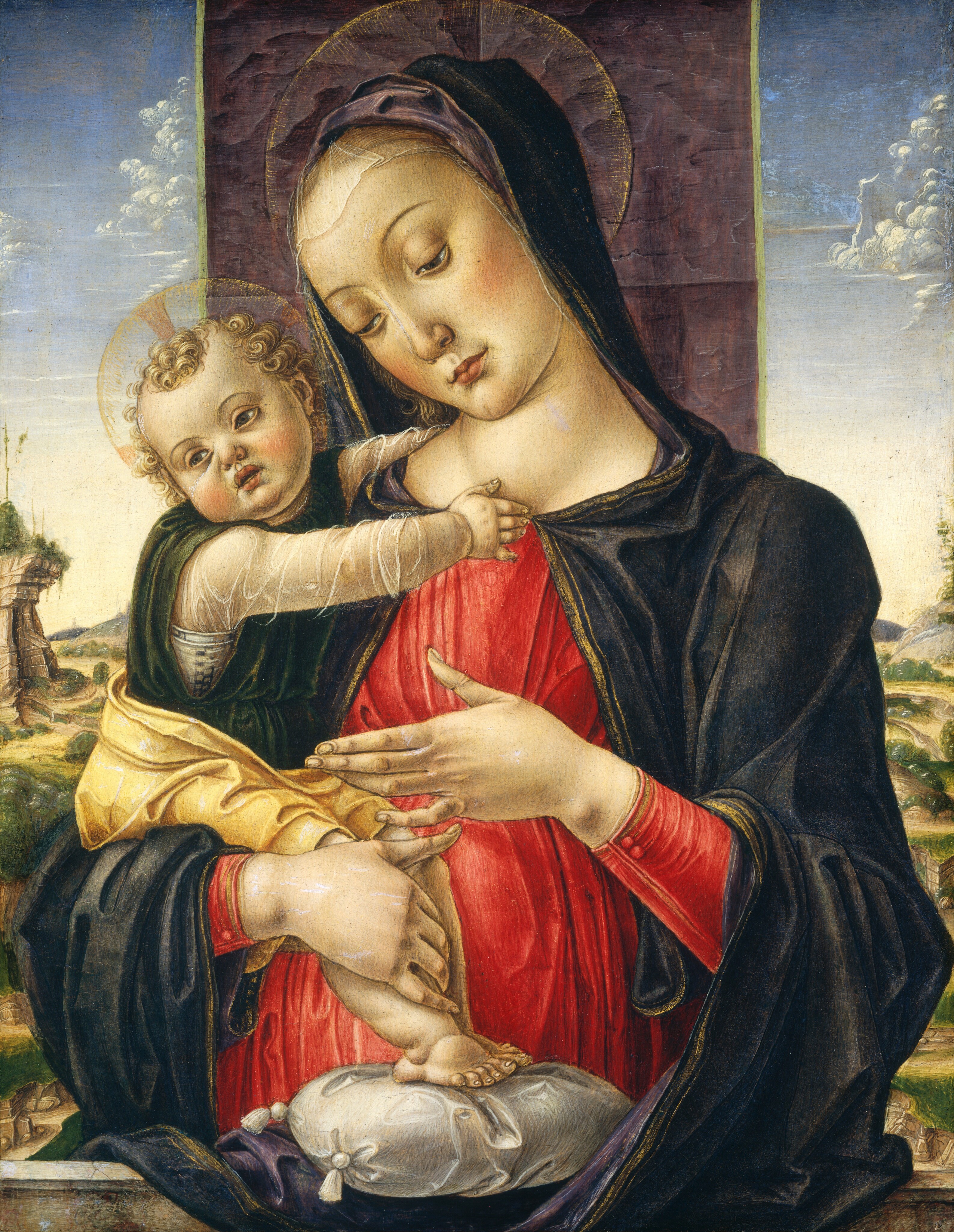 Shown from the waist up, a woman behind a stone ledge and in front of a landscape holds a chubby baby tucked into one elbow in this vertical painting. Both have pale skin, flushed cheeks, and blond hair surrounded by gold-edged halos. The woman looks down and to our left with hooded eyes. Her pink lips are closed over a pointed, cleft chin. A translucent white veil brushes across her high forehead and is covered by a marine-blue mantle that drapes over her head and shoulders. The mantle is edged in gold, and the plum-purple lining is visible where it is rolled back over her head and forearms. Her apple-red dress is cinched at the waist and falls in vertical folds. She holds her left hand, to our right, up with the palm facing in and the fingers gesturing toward the child. He half sits in the crook of her other elbow and rests his feet on a white pillow, which is on the gray stone ledge. The child wears a translucent white, long-sleeved garment under a pine-green tunic, and a butter-yellow cloth is wrapped around his hips. A purple banner hanging behind the woman is about the width of her collarbone, and a deep landscape to each side has trees, cliffs, and walking paths leading into the distance under piles of white clouds drifting across a watery blue sky.