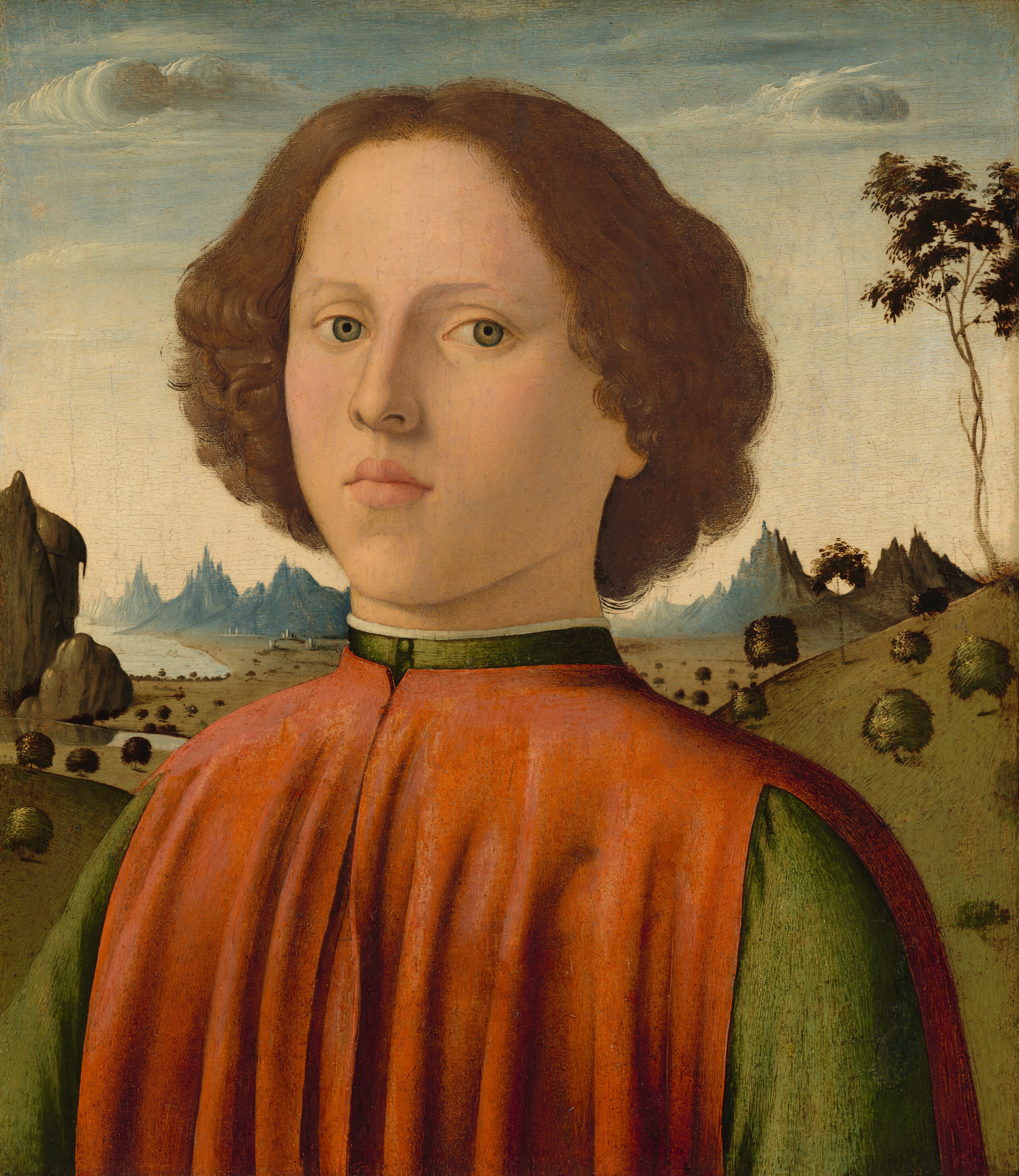 This painting is a portrait of a boy seen from the chest up, facing us with a neutral expression. He has fair skin, light eyebrows, defined, pink lips, and green eyes. His curly hair is brown and falls around his ears. The boy wears a high-collared garment with an olive-green undershirt and a reddish-orange outer vestment. There is a serene landscape in the background with rolling hills, pointed rock formations, scattered trees, bushes, and a blue sky with a few wispy clouds.