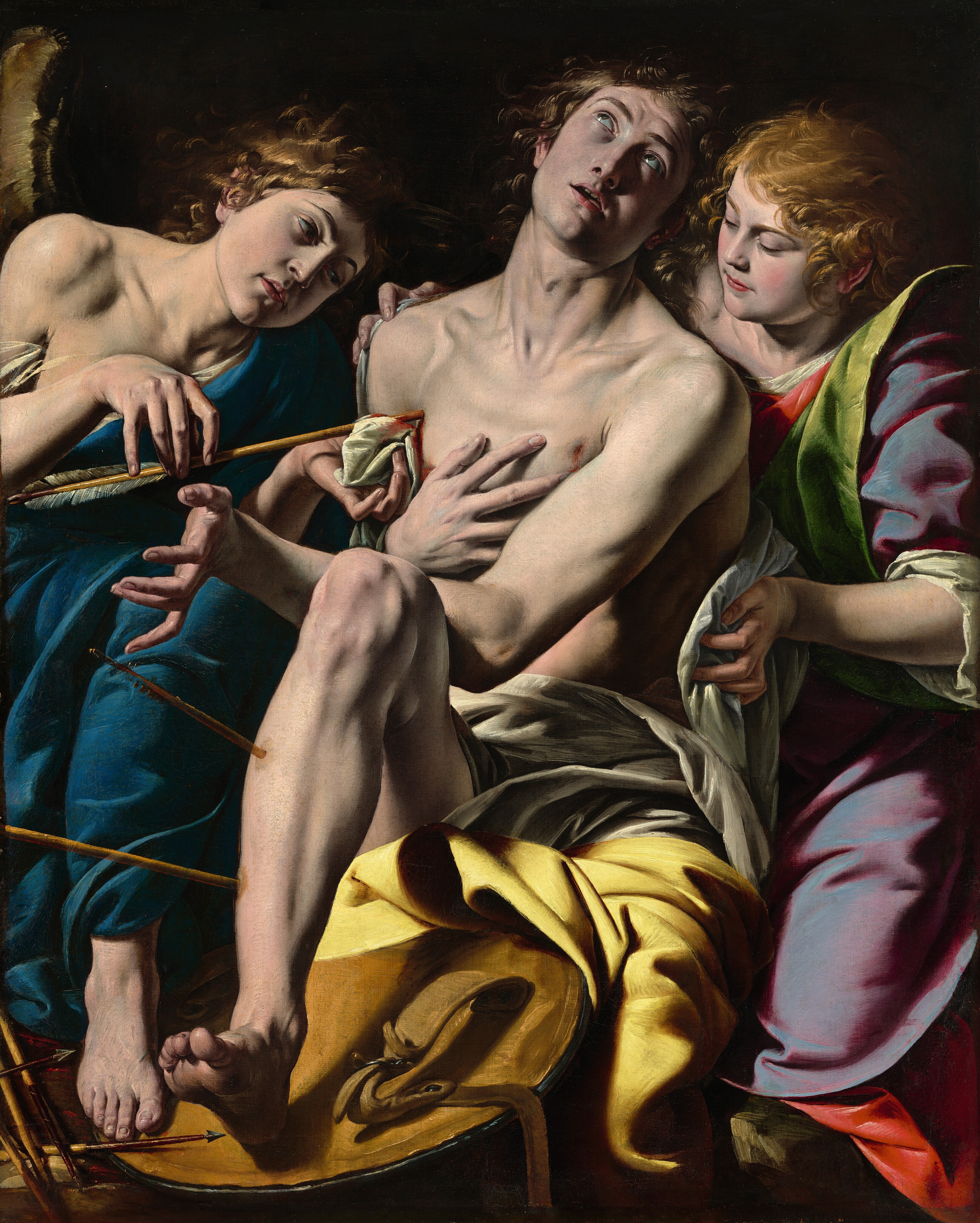 Bright light from the upper left washes over a young man pierced with arrows, an angel, and a young woman, who together fill this vertical painting. The three cluster closely against a black background, and all of them have pale, peachy skin. The muscular young man at the center, Saint Sebastian, faces us and appears to be seated with his right leg bent, to our left. The heel of that foot is braced on a round, golden brown object that might be the back of a shield. Saint Sebastian’s shin is pierced by two arrows. His head is thrown back as his glistening brown eyes roll back to gaze up and his mouth hangs open. He touches his chest near one wound, near his left shoulder, to our right. His other wrist rests against his bent knee, and the palm of that hand faces our left, fingers splayed back. He is nude except for silver-gray and goldenrod-yellow cloths draped across his lap and left leg. To our left, the angel, wearing a long, sapphire-blue toga, leans toward Saint Sebastian with shoulders squared to us. The angel’s head tips toward Saint Sebastian’s shoulder as he delicately draws an arrow from the saint’s chest and holds up a bloody cloth to staunch the wound. A sliver of a golden-brown wing shimmers against the ink-black background, near the left edge of the painting. Several blood-covered arrows sit near the angel’s feet, in the lower left corner of the painting. To our right, the woman stands close behind Saint Sebastian, wearing a plum-purple robe with emerald-green lining over a long, scarlet-red garment. She cradles Saint Sebastian’s body against hers, bracing him with her hands as she wraps the gray cloth around his back.