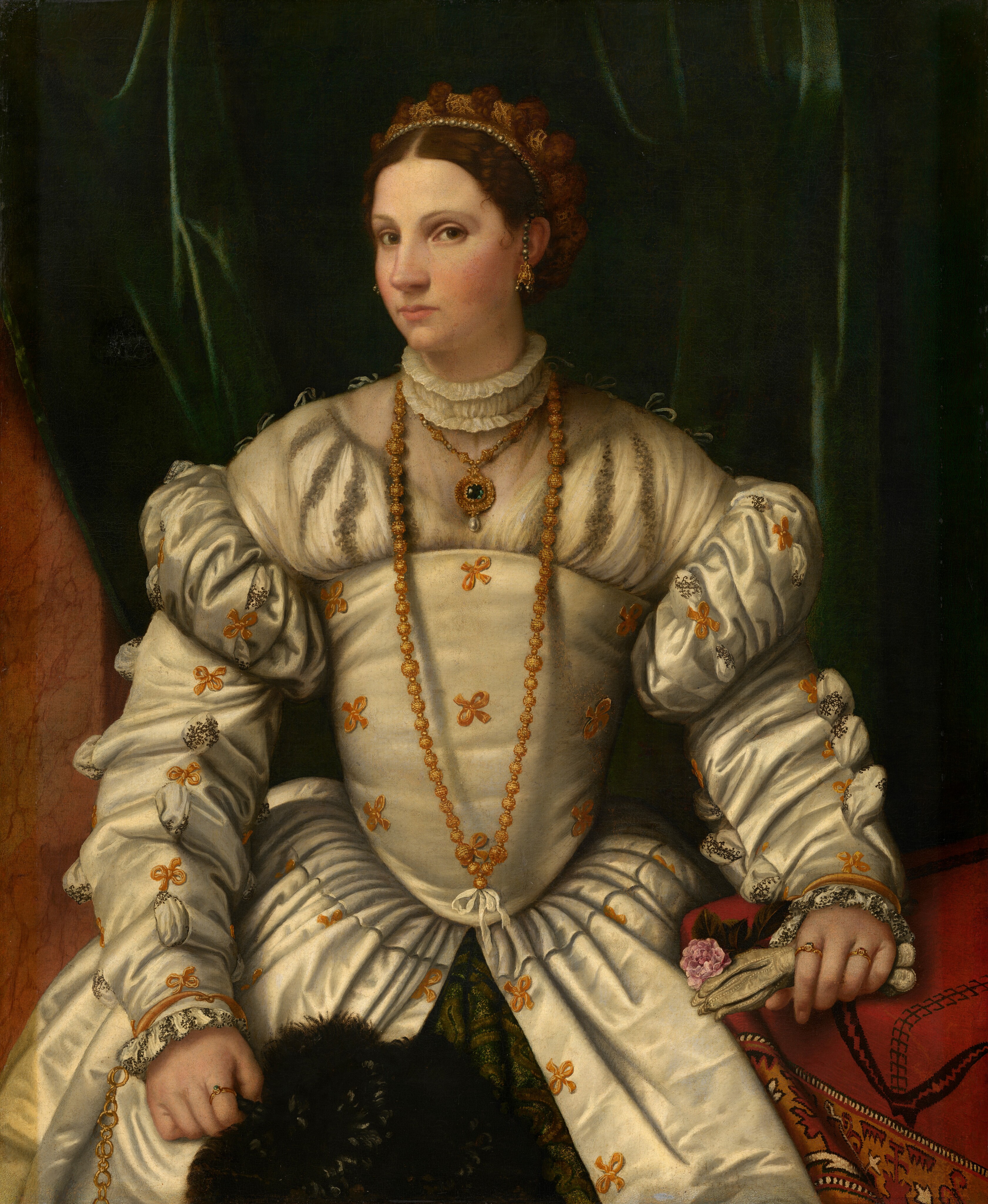 Shown from the thighs up, a pale-skinned woman wearing a cream-white gown ornamented with gold bows looks at us from the corners of her brown eyes in this vertical portrait. Her body faces us, but she turns her head to our left. She has thin, arched eyebrows, a straight nose, flushed cheeks, a dimple in her chin, and her pink lips are closed. Her chestnut-brown hair is parted down the middle and pulled back under a gold and pearl headdress, but two wispy curls lie on her smooth forehead near the temple we can see. A string of black pearls ending in a gold pendant hangs down the front of her ear. A white ruffled collar encircles her neck over two heavy, gold necklaces. A blue stone and a pearl in a gold setting lies on her chest, and the second chain reaches to her waist. The satin bodice is fitted, and the sleeves are puffy below the shoulders. Voluminous long sleeves are slitted and a white chemise patterned with delicate black designs is pulled through, creating little tufts. She wears three gold rings on her two hands. She holds white gloves and a pink carnation in one hand, which rests on a table covered in a red tapestry to our right, and a dark green feather fan in her other hand. Her gown splits over a pine-green skirt beneath. A shimmering, forest-green velvet curtain behind the woman sweeps to one side to reveal a caramel-grown stone pillar along the left edge of the painting.