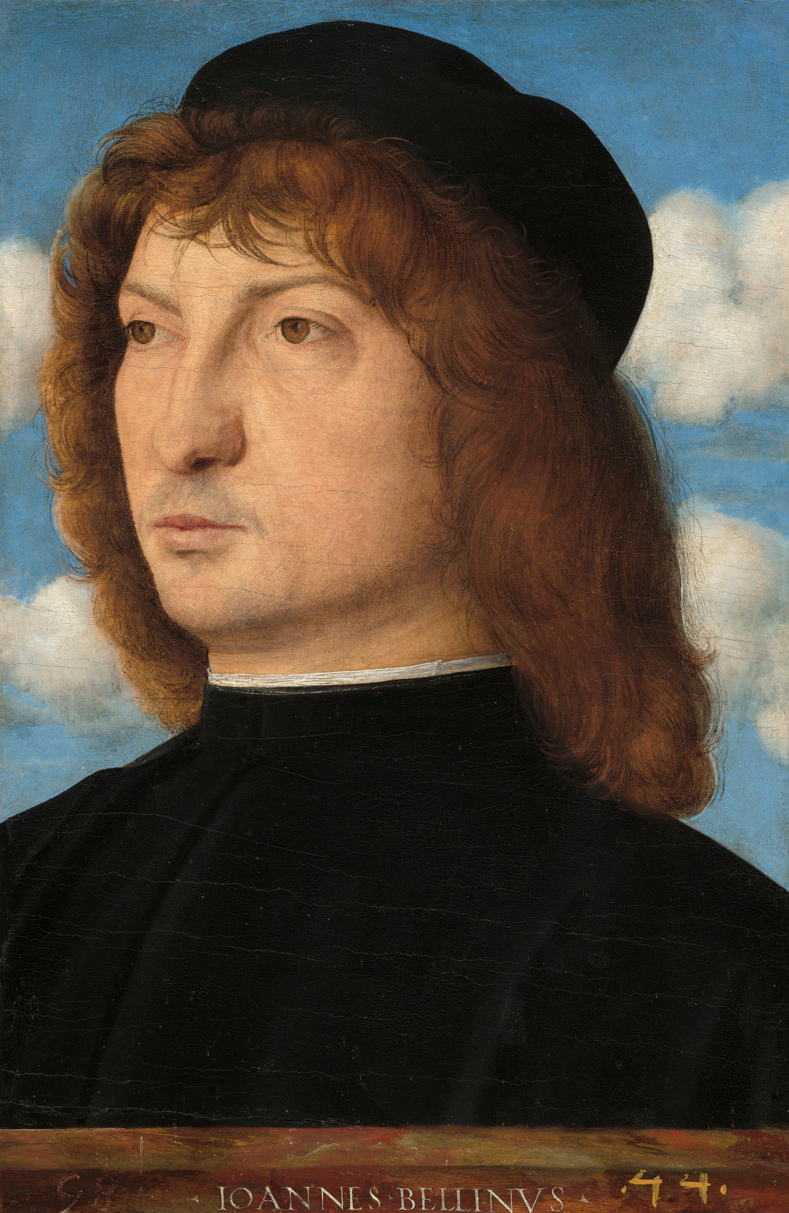 Shown from the shoulders up, a clean-shaven, pale-skinned man with shoulder-length brown hair and dressed in black fills this vertical portrait painting. His body is angled to our left, and his shoulders extend off both sides of the composition. He looks off to our left with brown eyes under thin brows. He has a long, slightly bumped nose and the hint of a five-o-clock shadow around his pale pink lips, which are closed. His brimless black cap nearly touches the top edge of the composition. His wavy hair is cut into bangs across his forehead and is feathered down each side of his face to meet the longer hair at the back. He has a high-necked, ink-black garment lined with white across his neck. Two bands of white clouds span the azure-blue sky behind him. A stone ledge in front of the man, across the bottom of composition, is painted to look like it had been carved with the artist’s name: “IOANNES BELLINVS.” Two numbers are painted in the lower corners, with “98” to the left and “44” to the right.