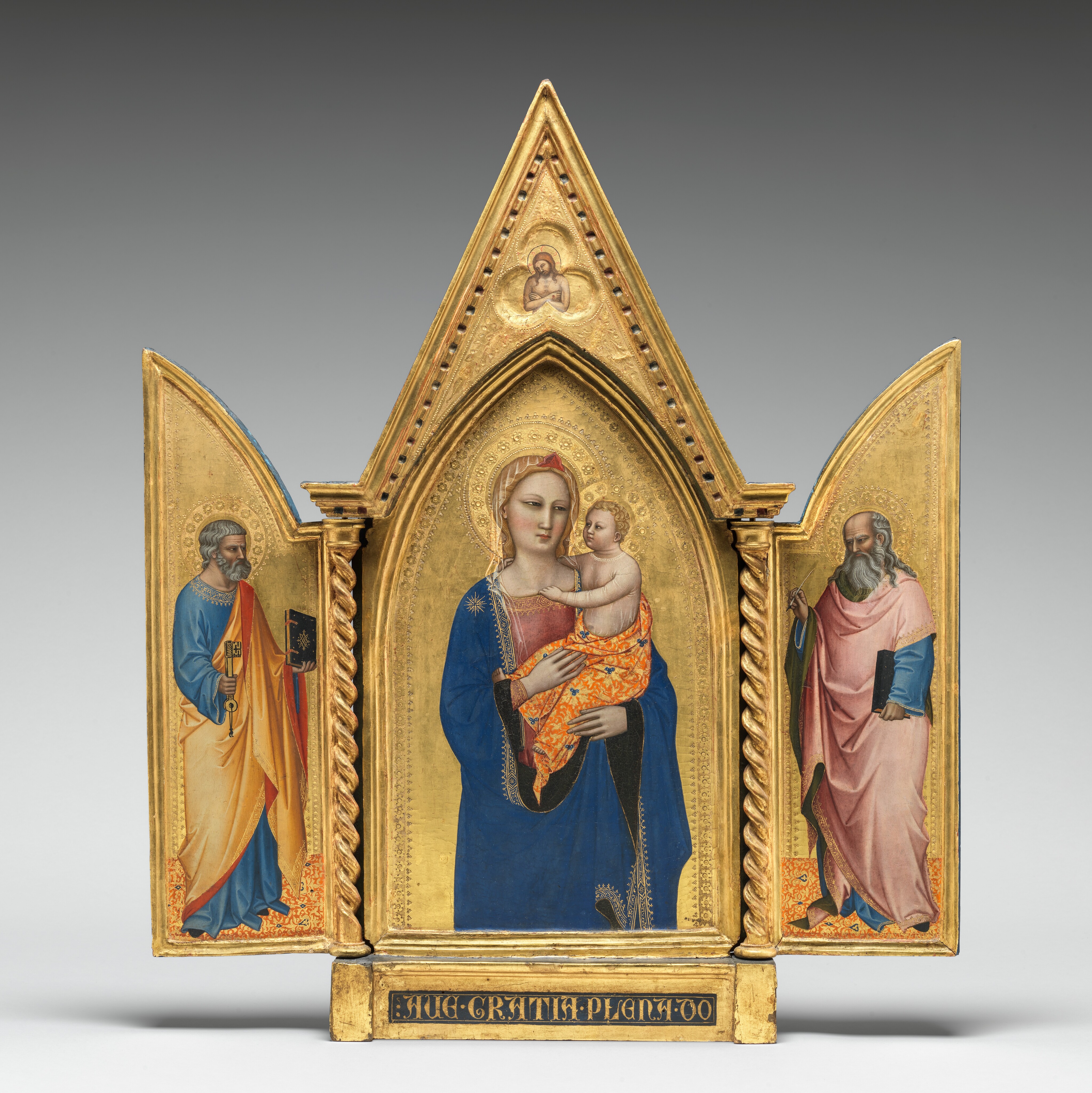 Two panels on a gilded, carved, wooden frame are swung open like shutters to show a bearded, gray-haired man wearing robes standing in each wing, on either side of panel with a pointed, arched top showing a woman holding a baby. In a three-lobed shape in the apex above the woman and child, a nude man crosses his arms over his chest. All the people have pale, ivory-colored skin. Gold halos encircle each person’s head and the background of all three panels is bright gold. The woman in the center panel is shown from the hips up with her body facing us. The deep blue mantle that drapes over her shoulders and her dusty rose-pink dress beneath are trimmed in gold. A sheer veil covers her wavy blond hair and a red, triangular form sits above her forehead like a diadem. She looks to our right with dark, almond-shaped eyes. She has a straight nose, her coral-pink lips are closed, and her smooth cheeks are lightly flushed with pale pink. She holds the child in the crook of her left arm, on our right, and rests her other hand in the baby’s lap. He has short, curly blond hair and looks directly at the woman. He reaches his right arm, farther from us, around his mother’s neck and hooks his other hand in the neckline of her dress. He wears a sheer shirt edged in gold, and the fabric draped around his hips is patterned with tangerine-orange and gold, with lapis-blue clover designs. He has rounded cheeks and a pudgy tummy, but his facial features and arms are scaled more like an adult. Twisted columns separate this central panel from the shutter-like panels to each side. A single person stands in each wing, filling most of the space, and both are angled and look towards the central panel. Both men have darker, olive-toned complexions, wear long robes trimmed in gold, and stand on floors patterned with orange, gold, and sparsely spaced royal blue diamond-shapes. The man to our left has short, steel-gray hair and a trimmed beard. Wrinkles line his forehead, and his brow is furrowed. He wears a topaz-blue, long sleeved garment under a honey-orange robe, the underside of which is ruby-red where it turns back over his wrists and shoulder. He holds a book with a dark cover up in his left hand, farther from us, and a large skeleton key in his other hand. In the right panel, a balding man has a fringe of long, gray hair and a flowing, full beard. His forehead is also creased with wrinkles and his brow is furrowed. The dark, olive-green lining of his rose-pink robe shows where it folds back at his neck, over his wrists, and down the front. His robe covers a topaz-blue garment as well. He holds a book, also with a dark cover, by his side with his left hand, closer to us, and holds up a feather quill with his opposite hand. The inner edges of each of the central panel and both wings are incised with bands of decorative floral and geometric designs punched into the gold backgrounds. In the pointed peak above the central panel, the nude man is shown from the waist up in a three-lobed, clover shape that would be visible even if the wings were closed. The man’s body faces us but his haloed head tips to his right, our left. He has a beard and long brown hair, and a hole dripping blood pierces the back of his left hand, visible on our right. The base of the tryptic is inscribed with gold letters in a dark field: “AVE GRATIA PLENA DO.”