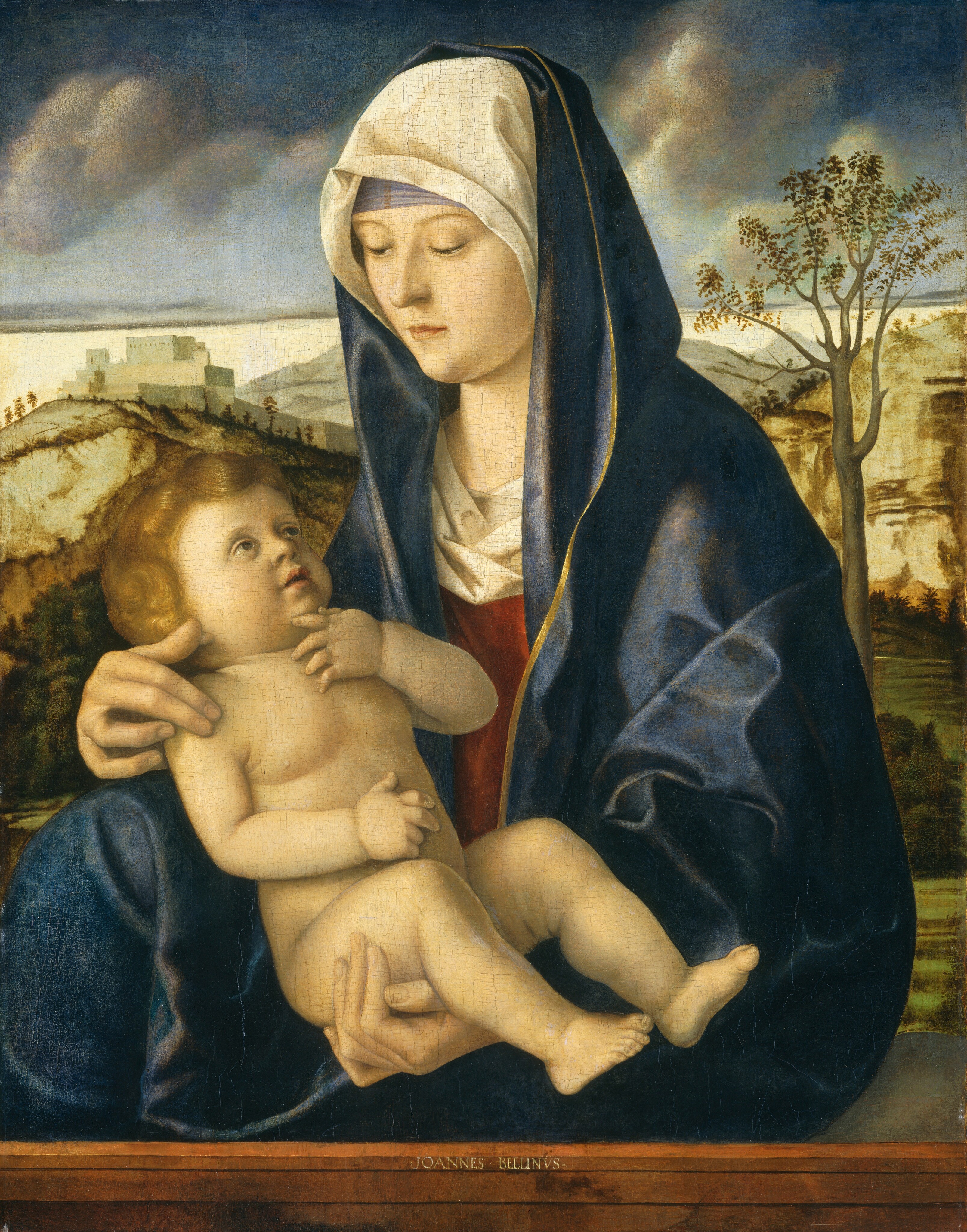 Shown from the hips up, a woman sits facing our left as she cradles a child in her lap in this vertical painting. A ginger-brown parapet spanning the lower edge of the composition separates them from us, and they have pale skin. A lavender cloth is wrapped across the woman’s forehead under an ivory-white veil that covers her hair. A cobalt-blue mantle drapes over her head and wraps around her brick-red robe. The mantle is edged in gold, and an ivory cloth is tucked into the neckline of the robe. Her eyes and head are lowered slightly to look at the child, and she supports his bottom with her left hand, on our right. Her right hand curves delicately around his shoulders. The nude, chubby child is cradled so he faces our right. He has delicate features, blond hair, and he looks  up to look at the woman with this lips parted. He rests one hand on his belly with the first two fingers raised, and he touches his chin with his other hand. Beyond the pair, a landscape is filled with grass, dark green shrubs, and tall trees. Hills climb to two distant, beige-colored hills streaked with dark brown and dotted with trees. A cluster of sage-green buildings stands on the hill to our left. Puffy clouds float in a dark azure-blue sky overhead. The brown stone parapet is inscribed in the lower center, “JOANNES BELLINVS.”