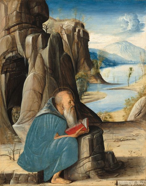 This painting depicts an old man sitting on rocky ground, holding a red book in his thin arms and facing the right. He has pale skin, a long white beard, and a balding head, and he is wearing a light blue hooded cloak that envelops his body. A thin golden halo surrounds his head, and his bare feet are visible on the rocky ground. On the left are tall, rocky gray mountains with a large dark cave. A lion-like animal is partially visible at the mouth of the cave, looking out towards us. On the left is an open landscape with a light blue river winding into the distance with sand-colored banks and tall white and blue mountains in the distance under a white and blue, partially cloudy sky.