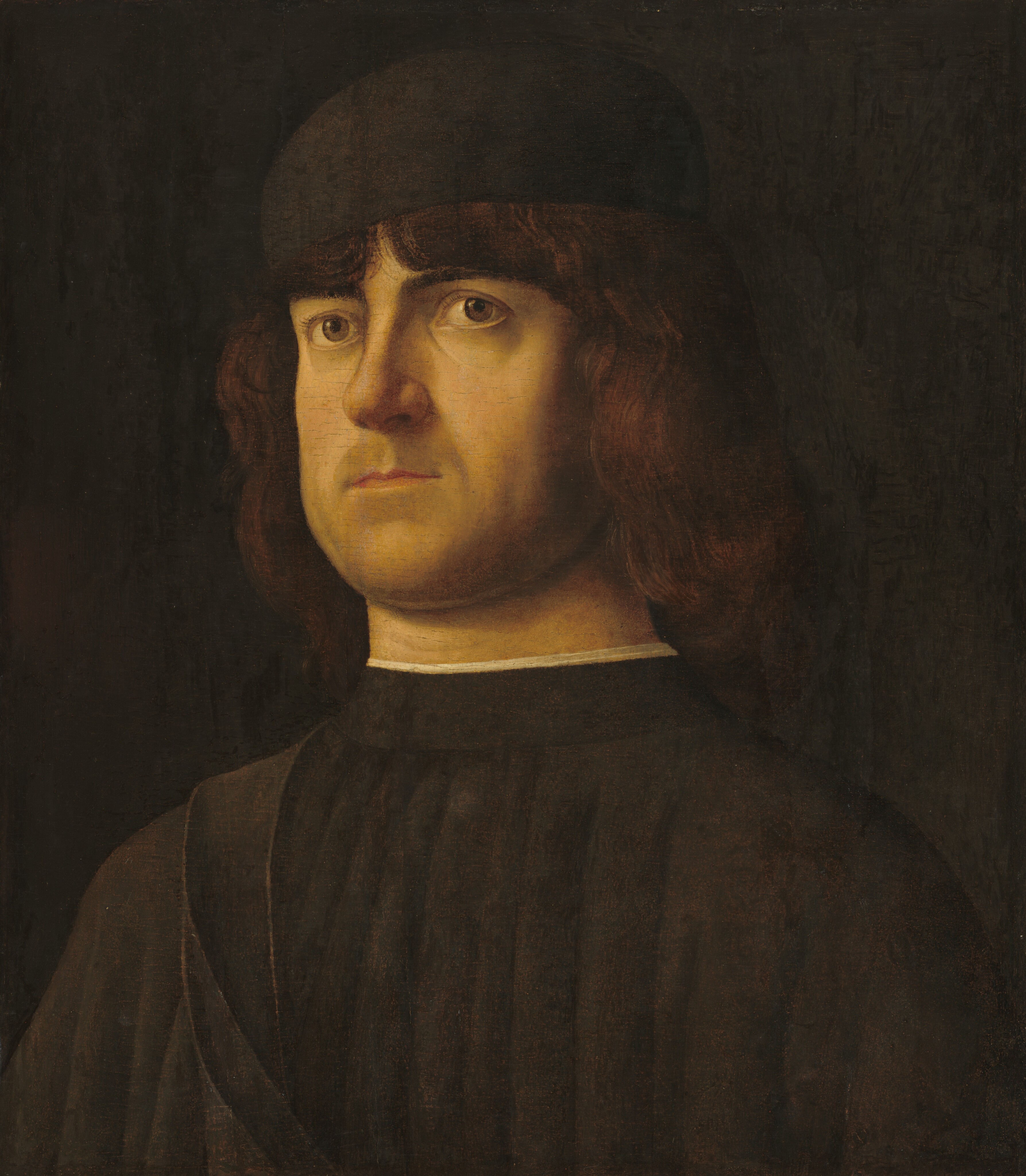 Shown from the chest up, a pale-skinned, clean-shaven man with brown hair and wearing brown looks up and slightly to our right in this vertical portrait painting. The man’s brown eyes gaze up under low, dark brows. He has a square face with a rounded nose, a wide chin, and his thin lips are closed. Wavy brown hair falls almost to his shoulders from under an earth-brown, brimless cap. Vertical pleats fall from the high neck of his coffee-brown garment, which is lined with a narrow band of white at the neck. The man is lit brightly from our left, but his clothing and hair nearly blend into the dark brown background.
