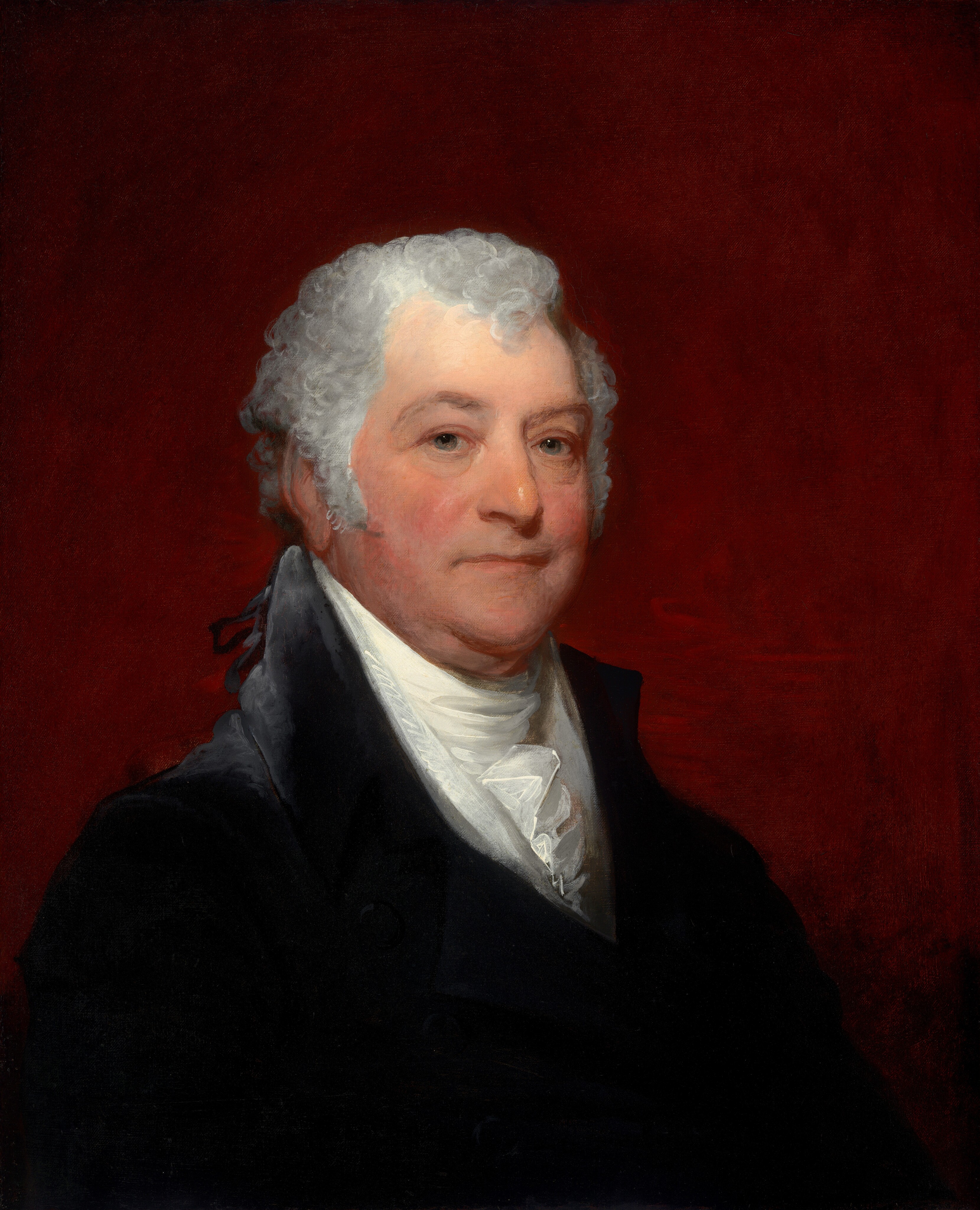 The image depicts the upper half of a man's body in a portrait. He is positioned slightly angled to his right but his face is turned towards the viewer. The man has a fair complexion with rosy cheeks, light-colored eyes, and a straight nose. His hair is white, curly, and close-cropped to his head. He is wearing a dark jacket with a high collar and a white cravat tucked into the jacket. The background is a solid deep red. There are no visible accessories or objects around him.