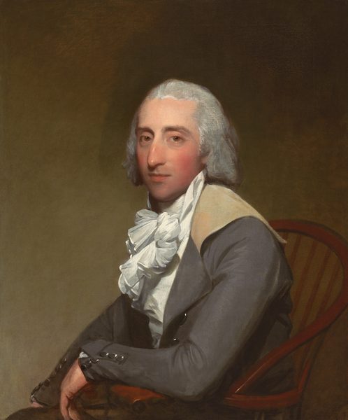 The image depicts a man seated in a wooden chair. He has light skin, gray hair, a prominent nose, and is wearing a formal outfit with a high-collared white shirt, a ruffled cravat, and a dark jacket. The man is facing the viewer directly with a slight smile. The background is plain and earthy brown in color.