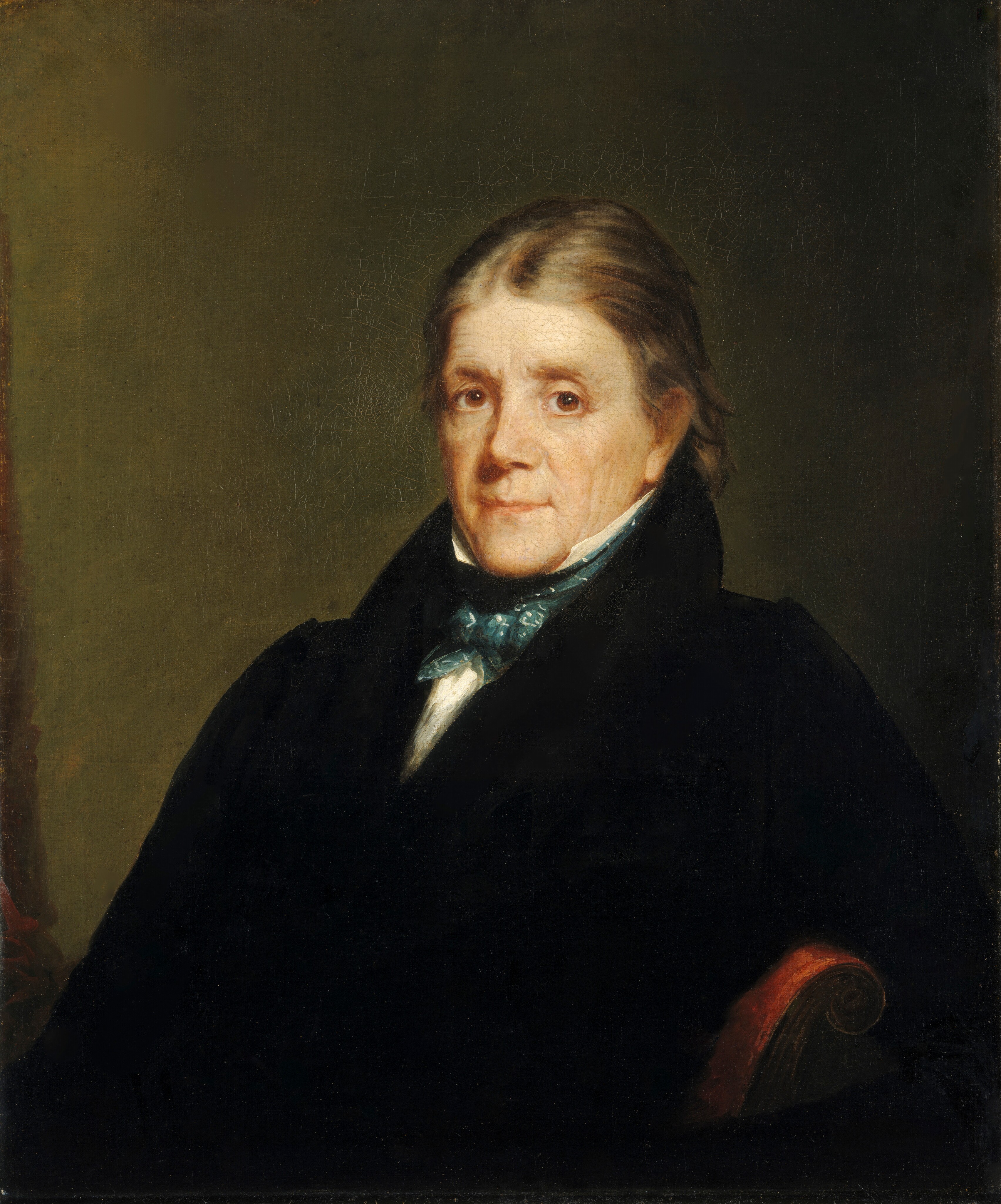 The image shows an elderly man seated, looking directly at the viewer. He has a gentle expression with deep-set eyes and gray hair combed back over his ears. He is wearing formal dark clothing with a white shirt and a blue cravat. There are no visible accessories. The background is a simple dark color.