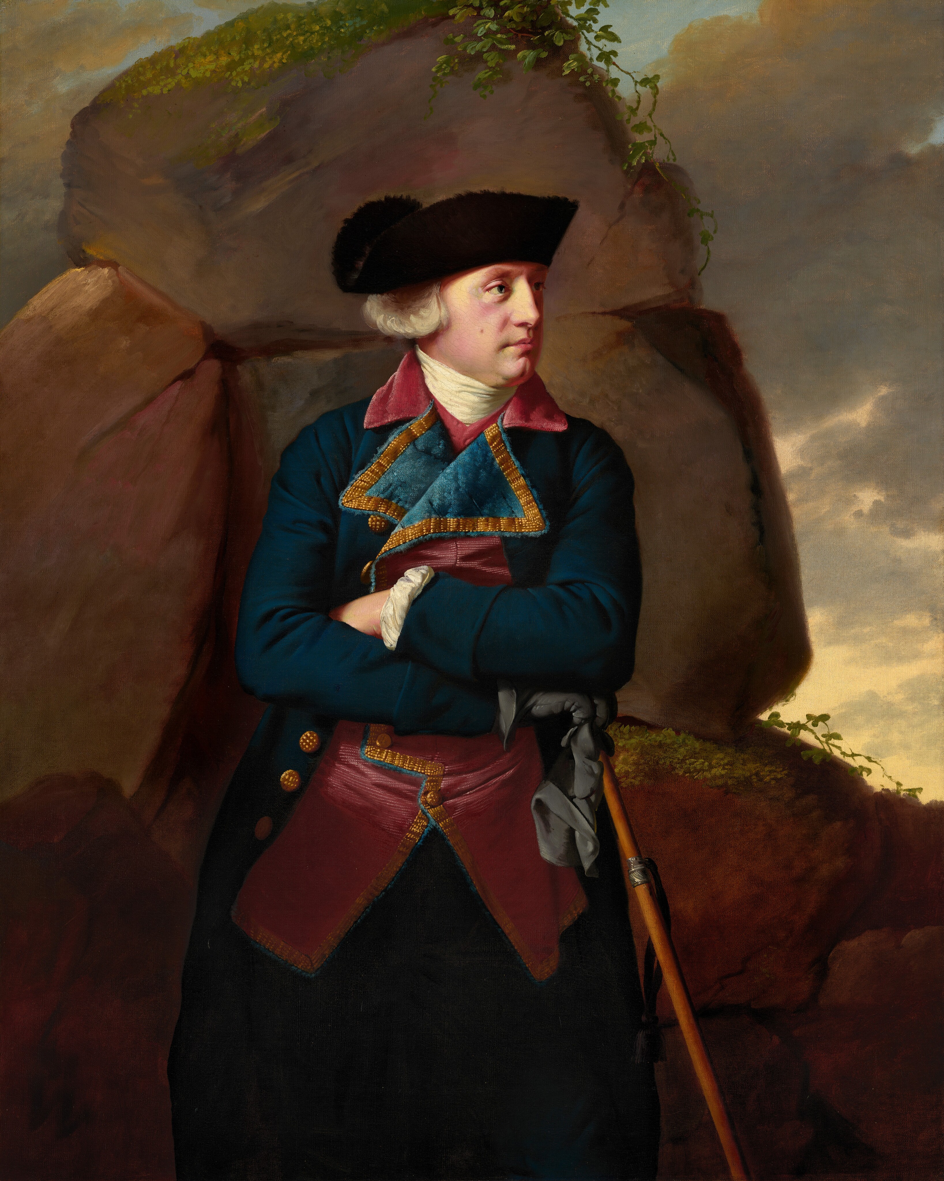 The painting depicts a man shown from the knees up, standing with his arms crossed over his chest and his head turned to the right. He has light skin, ruddy cheeks with a brown mole, a long nose, and small red lips in a neutral expression. His gray hair is styled into curls above his ears that peek out from under a black hat. His clothing is rich in detail, consisting of a dark blue-and-red coat with gold embroidery and gold buttons, and an off-white scarf or cravat wrapped around his neck. His right hand wears a black glove and holds a cane, as well as the other glove. The background features large, moss-covered rock formations, with a few green vines trailing across them. The pale-yellow sky behind it is filled with clouds, with some hints of blue at the top.