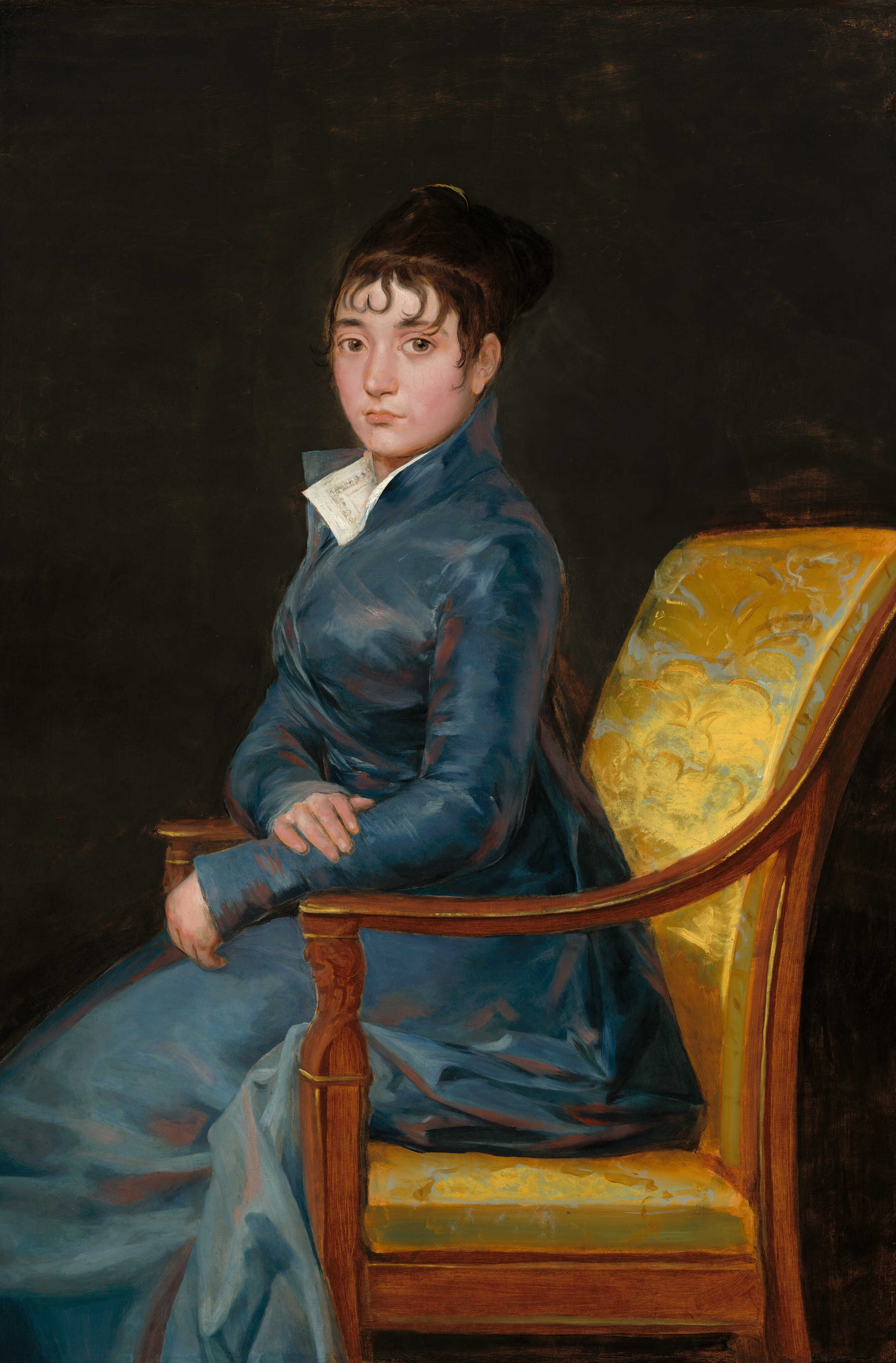 Shown from the knees up, a pale-skinned woman wearing a muted, sapphire-blue gown sits ramrod straight in a chair and looks at us in this vertical portrait painting. The chair and the woman’s body face our left in profile, but she turns her round face to look at us with large, light brown eyes. She has slightly flushed cheeks, a straight nose, a full lower lip, and her mouth is closed. Her left forearm, closer to us, rests on the front corner of the wooden arm of the chair. Her other hand lies across that forearm. Her blue dress has a high collar and wraps closed across her chest. The full skirt bunches around the back of the seat and around the arm of the chair. The corners of a white collar lie open at the woman’s throat, nestled into the dress's lapels. The dress is painted rather loosely with shades of slate and royal blue and shaded with mauve pink, giving it a hint of a sheen. The back and seat of the wood chair is upholstered in gold-colored fabric, which is also loosely painted with the suggestion of a brocade pattern.  The background is dark brown, nearly black.