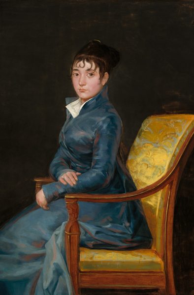 Shown from the knees up, a pale-skinned woman wearing a muted, sapphire-blue gown sits ramrod straight in a chair and looks at us in this vertical portrait painting. The chair and the woman’s body face our left in profile, but she turns her round face to look at us with large, light brown eyes. She has slightly flushed cheeks, a straight nose, a full lower lip, and her mouth is closed. Her left forearm, closer to us, rests on the front corner of the wooden arm of the chair. Her other hand lies across that forearm. Her blue dress has a high collar and wraps closed across her chest. The full skirt bunches around the back of the seat and around the arm of the chair. The corners of a white collar lie open at the woman’s throat, nestled into the dress's lapels. The dress is painted rather loosely with shades of slate and royal blue and shaded with mauve pink, giving it a hint of a sheen. The back and seat of the wood chair is upholstered in gold-colored fabric, which is also loosely painted with the suggestion of a brocade pattern.  The background is dark brown, nearly black.