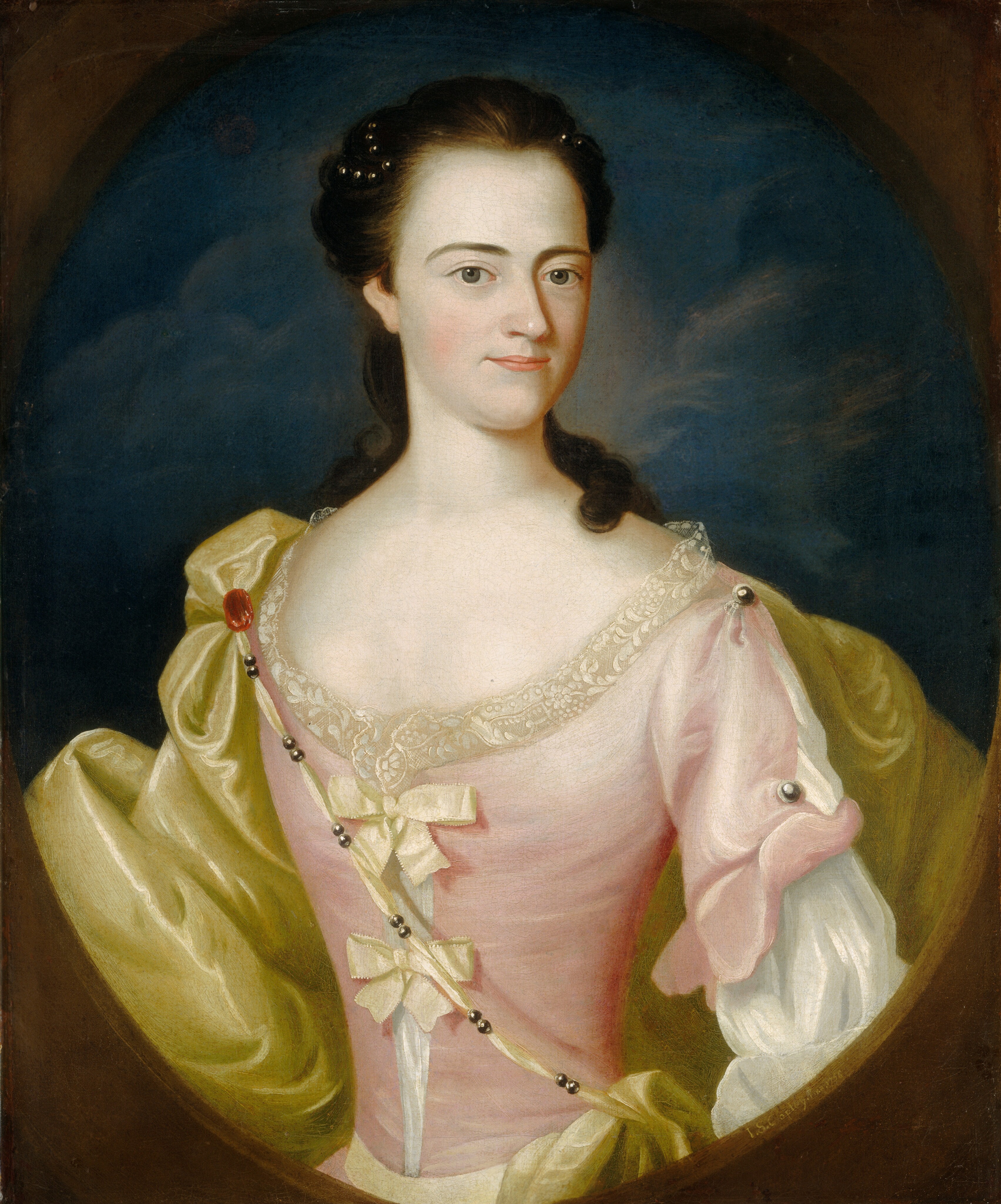 Shown from the waist up, a pale-skinned woman wearing a satin or silk shell-pink gown and a greenish-yellow cloak looks at us, slightly smiling, through an oval opening in this vertical portrait painting. Her shoulders angle slightly to our left, and her oval-shaped face turns slightly to the right. She cuts her dark gray eyes cut to the side to look at us from under arched brows. Her nose drops a little at the end, and her thin, pink lips are closed. Her dark brown hair is pulled up and woven with dark pearls reaching back from the temples. Curls fall down her back, and one rests over her left shoulder, to our right. The tightly fitting bodice has low curved neckline lined with lace. Short sleeves embellished with pewter-gray buttons or bosses overlap longer white sleeves. The bodice is open down the front to show the white undergarment and is tied with two pale yellow bows, one over the other. The cloak falls heavily in deep folds from her shoulders and is held in place with a strip of fabric lined at intervals with pairs of dark silver beads. The ribbon is affixed to the cloak with a red brooch at the woman’s right shoulder, to our left, and it stretches down and across her body. She stands in front of a dark blue, cloudy sky. The painted frame through which she looks is camel brown. The artist signed the front-facing, beveled edge of the oval frame: “I.S. Copley. Pinx. 1756.”