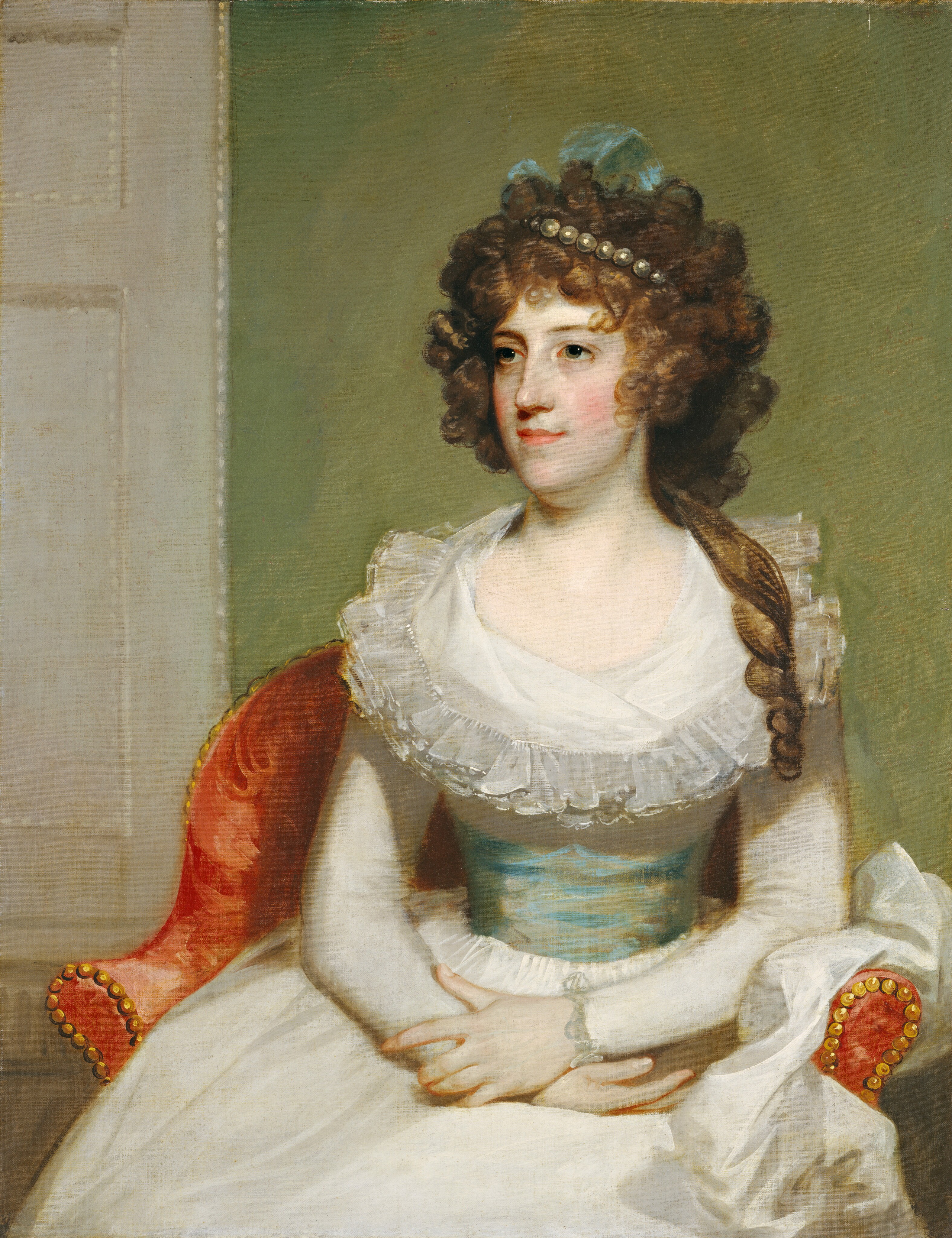 This painting is a portrait of a woman. She is seated on a red chair with gold studs along its edges, positioned with her arms crossed over her lap, slightly turned to the left. The woman's facial features include a fair complexion with rosy cheeks, slightly smiling lips, and large, dark eyes. Her brown hair is curly and voluminous, adorned with a pearl band and a light blue ribbon visible towards the back. Her clothing consists of a long-sleeved white dress with a ruffled neckline, cinched in the waist by a light greenish-blue sash. The background is a plain, muted green tone, with a light color on the left indicating a partially visible door or paneling.
