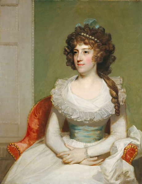 This painting is a portrait of a woman. She is seated on a red chair with gold studs along its edges, positioned with her arms crossed over her lap, slightly turned to the left. The woman's facial features include a fair complexion with rosy cheeks, slightly smiling lips, and large, dark eyes. Her brown hair is curly and voluminous, adorned with a pearl band and a light blue ribbon visible towards the back. Her clothing consists of a long-sleeved white dress with a ruffled neckline, cinched in the waist by a light greenish-blue sash. The background is a plain, muted green tone, with a light color on the left indicating a partially visible door or paneling.