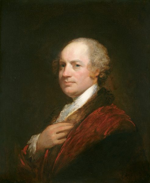 The image shows a portrait of a man's upper body. He is seated or standing, looking slightly to his right. The man has a round face, fair skin, prominent eyes, and light-brown curly hair. He is wearing an elegant red garment with fur trim around the collar. One of his hands is touching his clothing at the chest. The background is a dark, neutral shade, emphasizing the man's features and attire.