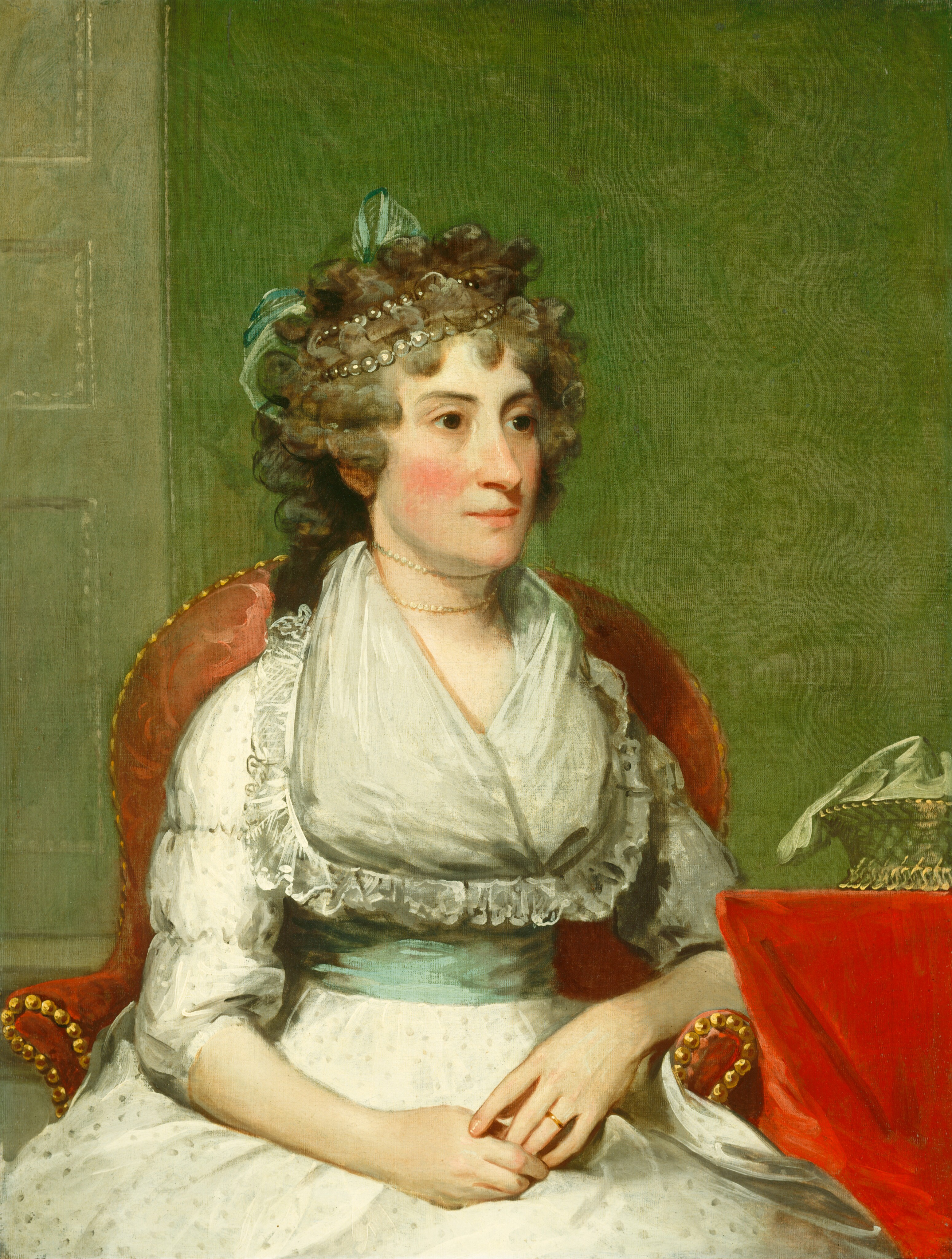 This is a painting of a woman depicted from the waist up. She is seated in a red armchair with gold details, slightly turned to the right, with her hands resting in her lap. She has light skin and slightly flushed cheeks, and her gray hair is styled in curls and adorned with a pearl headpiece and a light green ribbon. The woman is wearing a pale-green dress with a high neckline and a light blue sash around her waist, as well as lace trim on her neckline and sleeves. She has a gold ring on her finger and a thin necklace around her neck. Beside her on the right is a table covered with red cloth, with a small basket draped in a light-colored fabric on top of it. The background of the painting is a muted green wall with a door partially visible on the left.