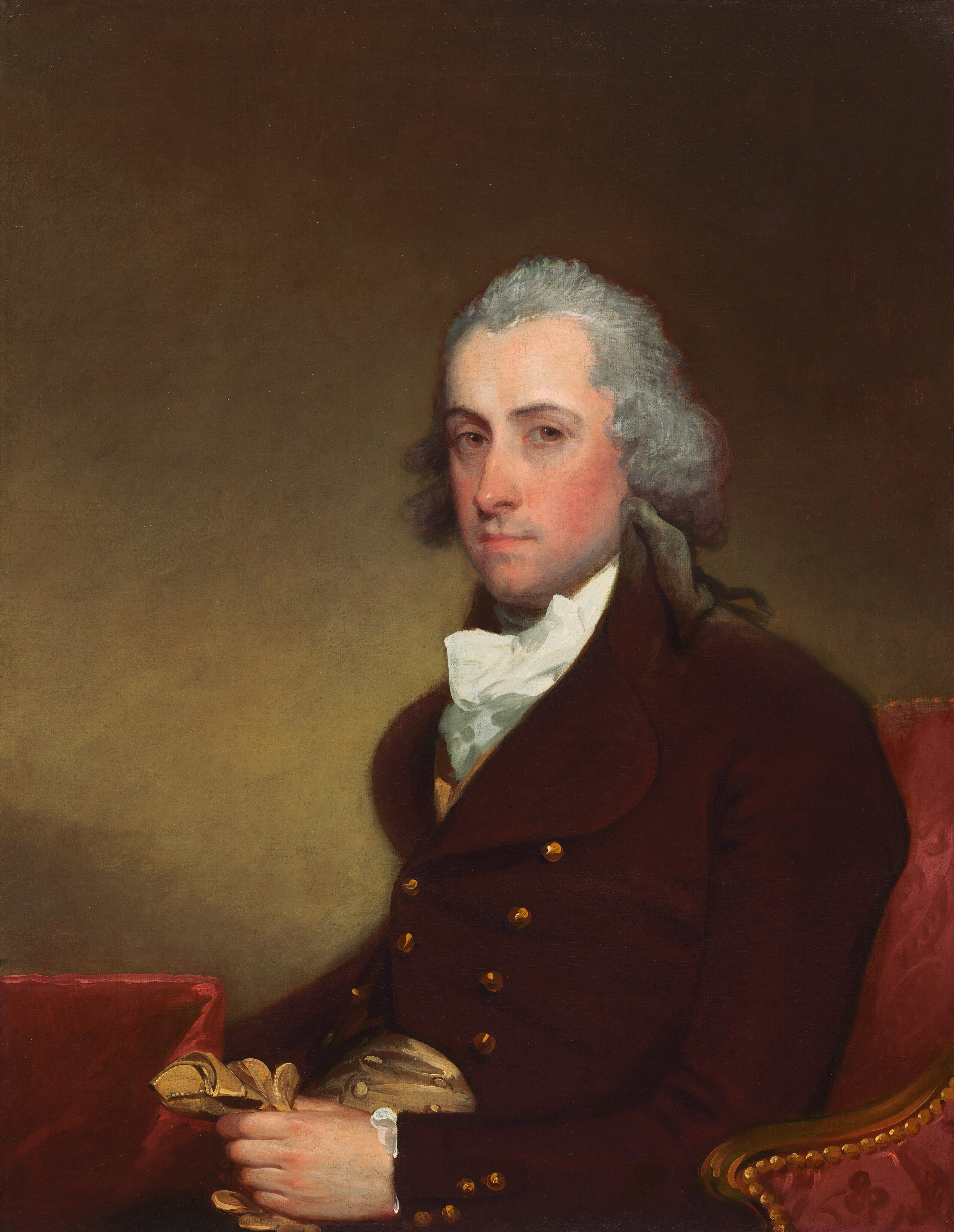 This painting is a portrait of a man. He is seated on a red cushioned chair, turned slightly to the left, facing us. He has a pale complexion and a neutral expression, with dark eyebrows, rosy cheeks, and thin lips. His hair is gray, with curls falling to the sides around his ears. He is dressed in formal attire, wearing a dark red, double-breasted coat with large golden button and a white cravat around his neck. The man holds a pair of tan gloves in his left hand, and in front of him is a piece of red furniture covered with a red cloth. The background is a muted brown gradient.