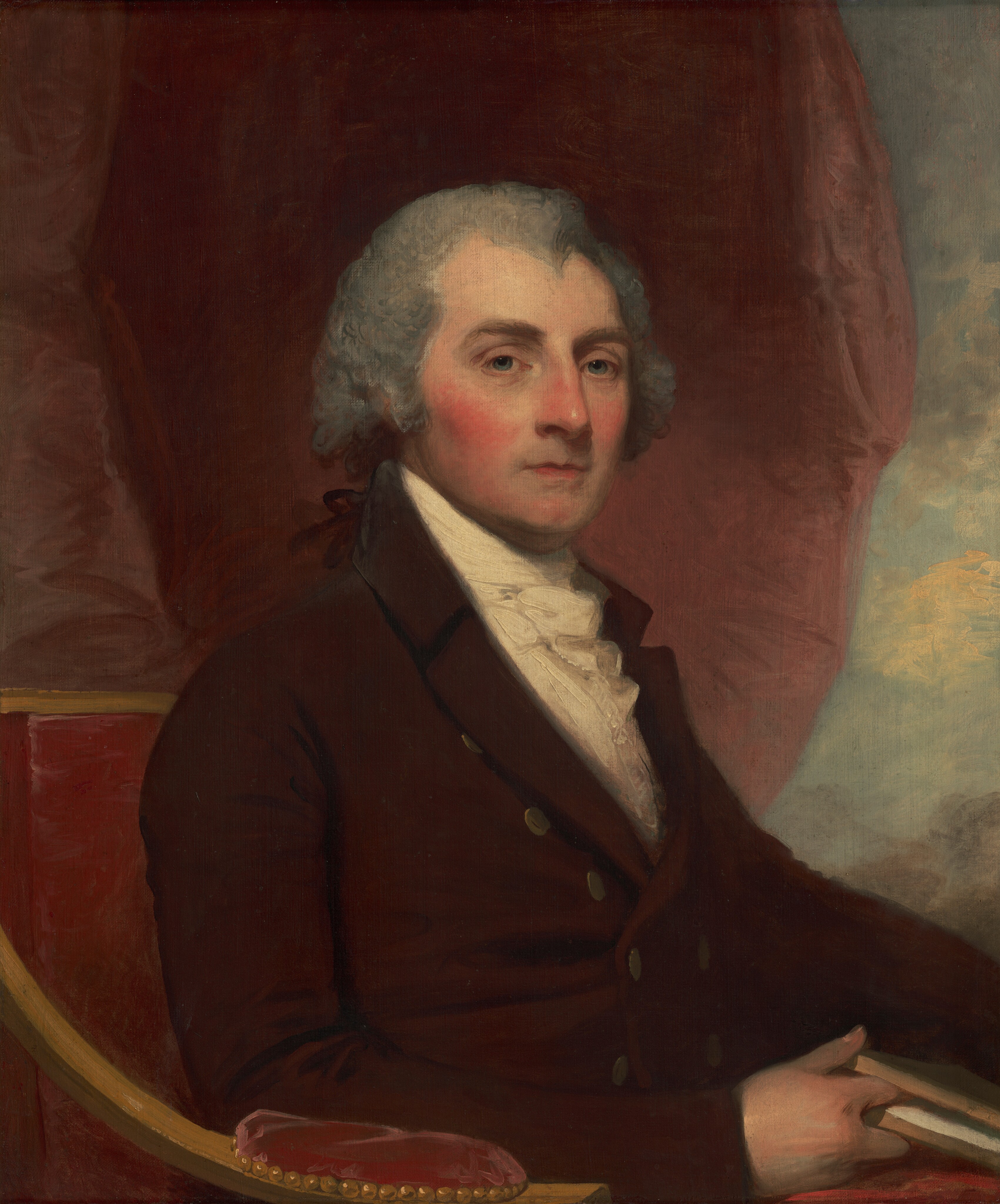 This painting is a portrait of a seated man. His body is facing the right with his head turned to us, a neutral expression on his face. The man has pale skin, rosy cheeks, bright blue eyes, thin lips, and gray hair that curls around his ears. He is dressed formally in a dark red coat with gold buttons and a white cravat at his neck, sitting in a red upholstered chair, holding a book in his right hand. The background consists of red drapery on one side and blue and yellow hues on the other, suggesting a cloudy sky.