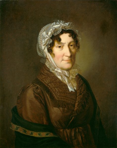 The portrait shows the upper half of a seated woman with a round face, wrinkles, and dark hair. She wears a bonnet, a brown dress with layers, and a shawl or cloak. The background is a muted dark brown.