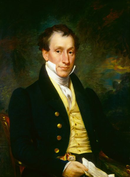 This image depicts a man from the waist up. He is seated, slightly turned to his right, while facing the viewer. The man has a composed expression, prominent cheekbones, and a slightly pointed nose. His dark brown hair is styled in a classic early 19th-century manner, with curls around the sides and forehead. He is dressed in a dark blue coat with gold buttons, a yellow waistcoat, a high-collared white shirt, and a decorative pin. In his right hand, he holds a piece of folded paper with writing on it. The background is an indistinct swirl of darker colors.