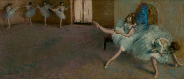 In a dance studio, two ballerinas wearing knee-length tutus and ballet slippers sit on a bench and stretch to our right while four more pose at the back of the space, to our left, in this long, horizontal painting. The scene and dancers are loosely painted so many features are indistinct. All the women have light skin and dark brown hair. Each pale, robins’ egg-blue costume has a low-cut, tightly fitting, sleeveless bodice and a full skirt. Dabs of off white, teal, and blue suggest flowers lining the deep U of the neckline at the front and down the back of each bodice. Closest to us and nearly brushing the edges of the lower right corner of the canvas, one dancer sits on the end of a wooden bench and leans down between her legs, her feet widely planted. She rests her torso on an elbow folded onto one knee as she reaches for her left foot, farther from us, with her other hand. Her hair is pulled up and her face, looking downward, is deep in shadow. Sitting behind her on the bench, to our left, a second dancer lifts her right leg, toe pointing, as she pulls on her pale pink tights. Her other leg stretches straight, pointed toe brushing the floor, in front of her. This woman’s hair hangs loose around her shoulders. The pair is brightly lit from a source to our right, and an arched area of electric blue and mustard yellow behind them is difficult to interpret. The floor, which takes up about two-thirds of the composition, is painted with dabs of teal blue, pale fuchsia pink, and caramel brown. It leads us back to the upper left corner of the painting where four more four ballerinas stand in a line. The pair to our left stands with their backs to us, facing the wall with their right legs raised to shoulder height, toes pointed. The pair to our right faces us. They also stand with their right legs raised, creating a wide angle with their standing legs. All four ballerinas bend their arms gently at the elbow, and some tip their heads toward the raised legs. The rightmost dancer is illuminated by a pair of French doors at the back corner, at the top center of the painting. The wall behind the bench is streaked with mint green over honey brown, and the wall at the back of the room is leather brown. The visible, soft brushstrokes give the image a blurred, ethereal appearance in the dimmed studio. The artist signed the painting in the lower left corner, “Degas.”