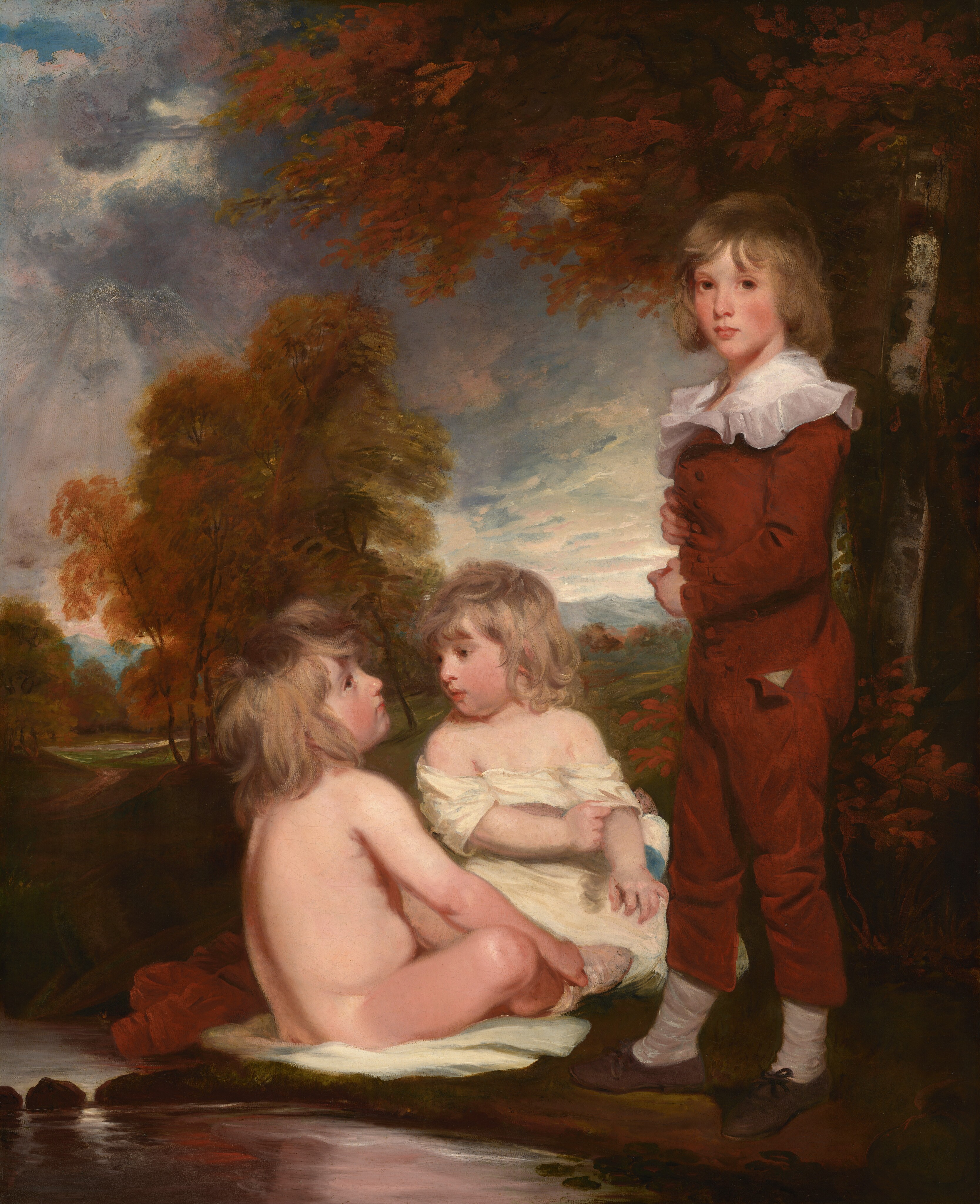 This painting shows three children in a natural setting. The child on the right is standing, visible from head to toe, while the two children on the left are seated in the grass. The standing child is positioned slightly turned to the left, with their arms folded across their chest. They have light brown, wavy hair that falls around their face, which has soft features and light complexion. This child is dressed in a russet-colored outfit with a ruffled white collar, white socks, and dark shoes. The seated child on the far left faces toward the standing child and has long, tousled, light-brown hair and a fair complexion. They are nude and sitting on a white cloth draped over the grass. The other seated child, sitting between the other two with their head turned to the left, wears a light garment partially draped over their shoulders and also has wavy, light-brown hair. The background includes trees with fall-colored foliage, a reflective stream running beside the children, and distant, cloudy skies.