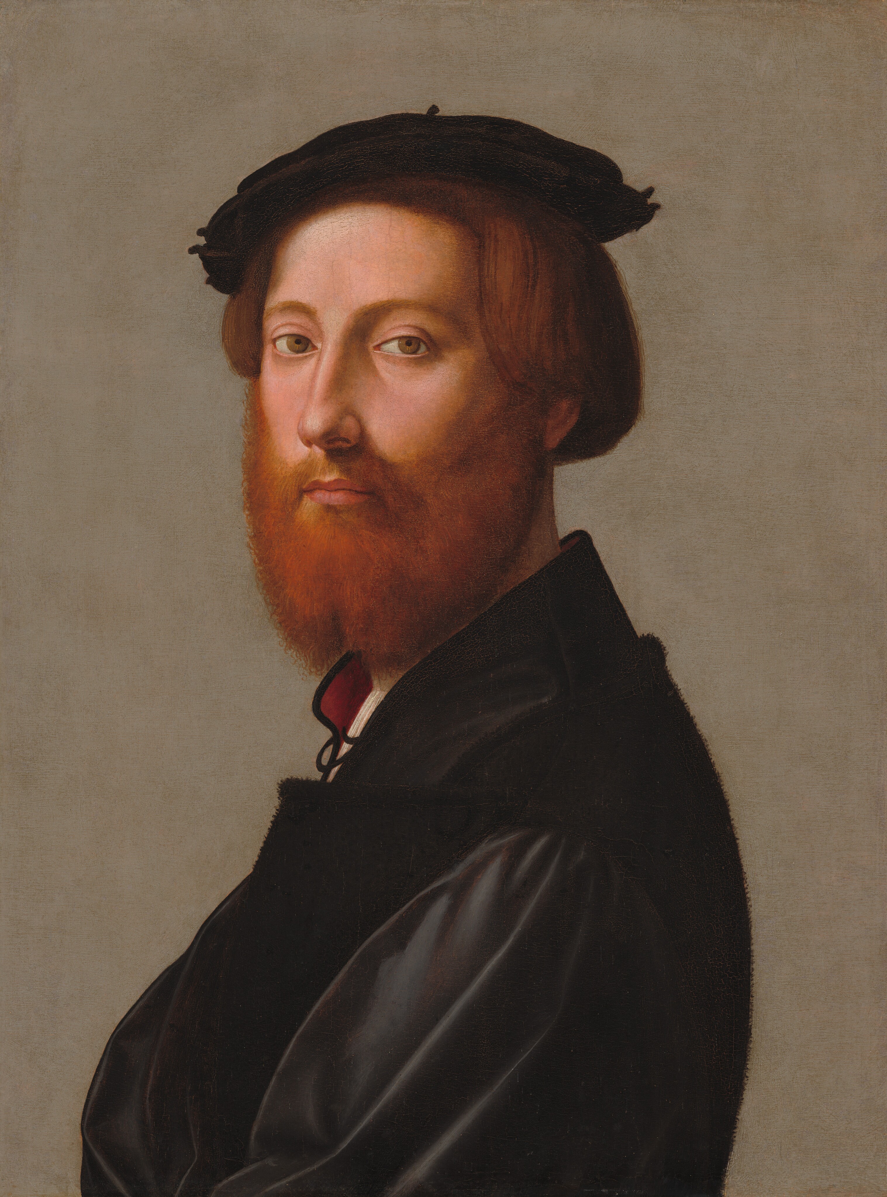 This painting is a portrait of a man depicted from the chest up, his body in profile, facing left, with his head turned toward us. He has pale, pink-toned skin, a full reddish-brown beard, short brown hair, arched eyebrows, a long nose, and thin pink lips in a neutral expression. He is wearing a black coat with a white garment and red collar peeking out by his neck. He also has a black beret or hat on his head. The background is muted gray.