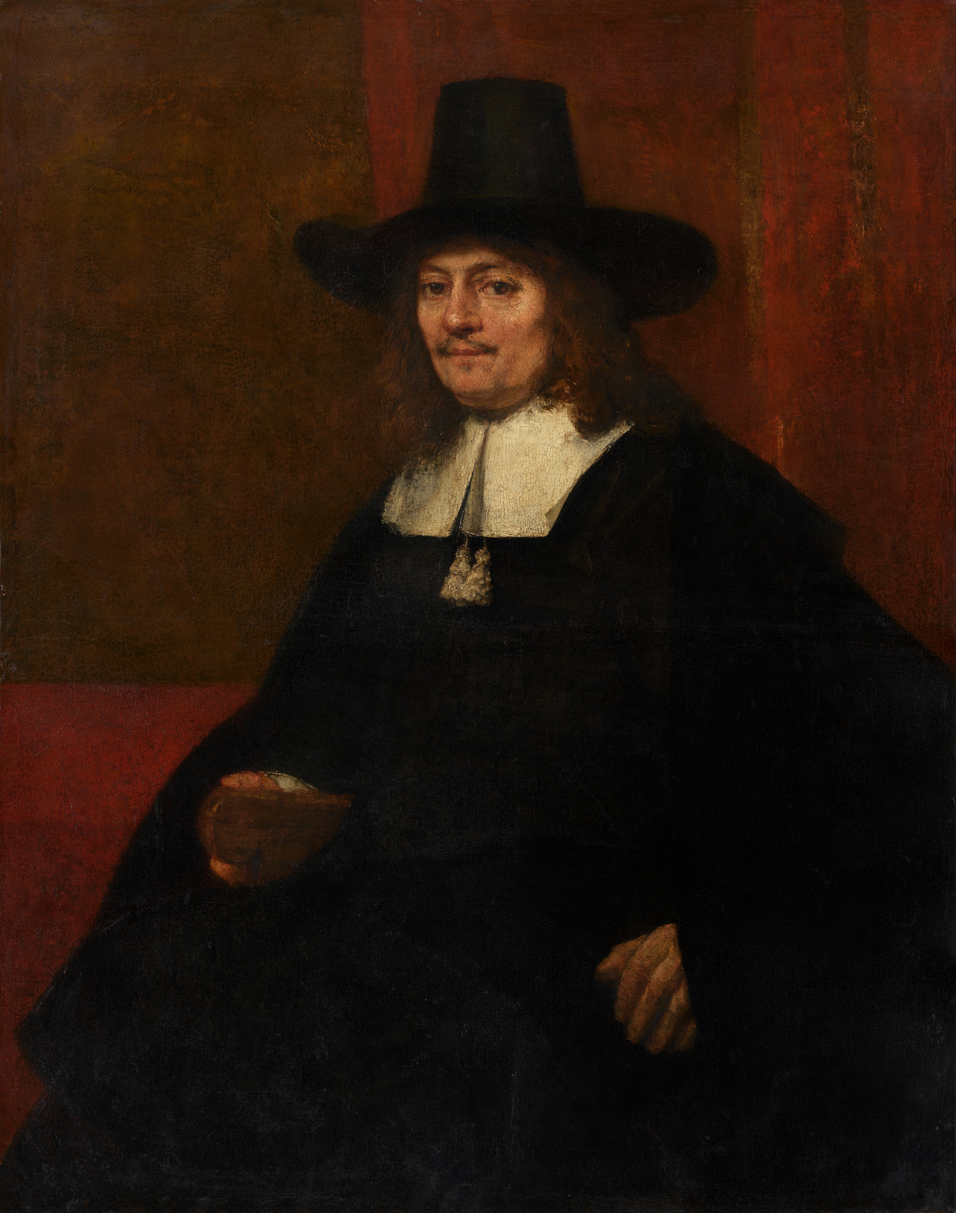 Shown from about the knees up, a man with peachy skin, wearing a voluminous black cloak and a tall, brimmed hat, looks directly at us in this vertical portrait painting. His body is angled to our left and he turns his face to look at us with brown eyes. He has a wispy, brown mustache over his pale pink lips, which are closed. He has a slight double chin and his reddish-brown hair falls to his shoulders. His wide, brimmed hat has a tall crown, which tapers a bit near the top. His black cloak falls over his arms and body, and has a wide, flat, white collar. Two white tassels hang below the split down the front of the collar. His left hand, to our right, is braced against the arm of a chair, and he rests his hand around the wooden end of the chair's arm. The wall behind him is brick-red with a coffee-brown rectangle to our left. Two vertical, fawn-brown stripes run down the red wall to our right, behind the man. Brushstrokes are visible in some areas, as in the man’s face, and a network of cracks runs across the surface of the canvas.