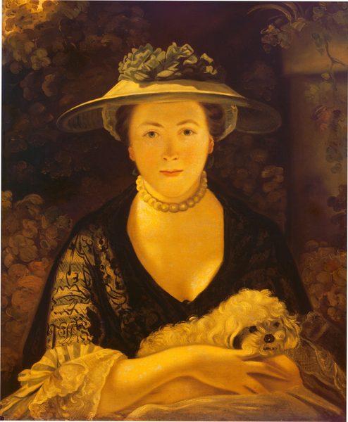 Shown from the lap up and shaded entirely in tones of golden yellow, laurel green, and black, a pale-skinned woman cradles a small, furry dog in her arms in this vertical portrait. The woman’s body is squared toward us, and she looks directly out with slate-blue eyes. She has an oval face with a delicate nose, long chin, and her pale pink lips are closed in a slight smile. Her brown hair is pulled back under a tan-colored hat lined on the underside with translucent cloth and on the top with sage-green ribbons or ruffles. A choker of thumbnail-sized pearls encircles her neck. Her dress has a deep V in the front and the elbow-length sleeves end with layers of lacy ruffles. Curving black lines suggest a pattern on sheer black fabric that covers pale yellow and frosty green stripes of the bodice. The dog has long, light-colored fur and a dark, bead-like nose and eyes. Vines grow up and from the top of a stone column in the background to the right, and the rest of the shallow space is filled with shrubs and plants. A bright yellow spot near the top left corner suggests a strong light source coming from that area, which casts her face in a diffuse shadow under the hat’s brim.