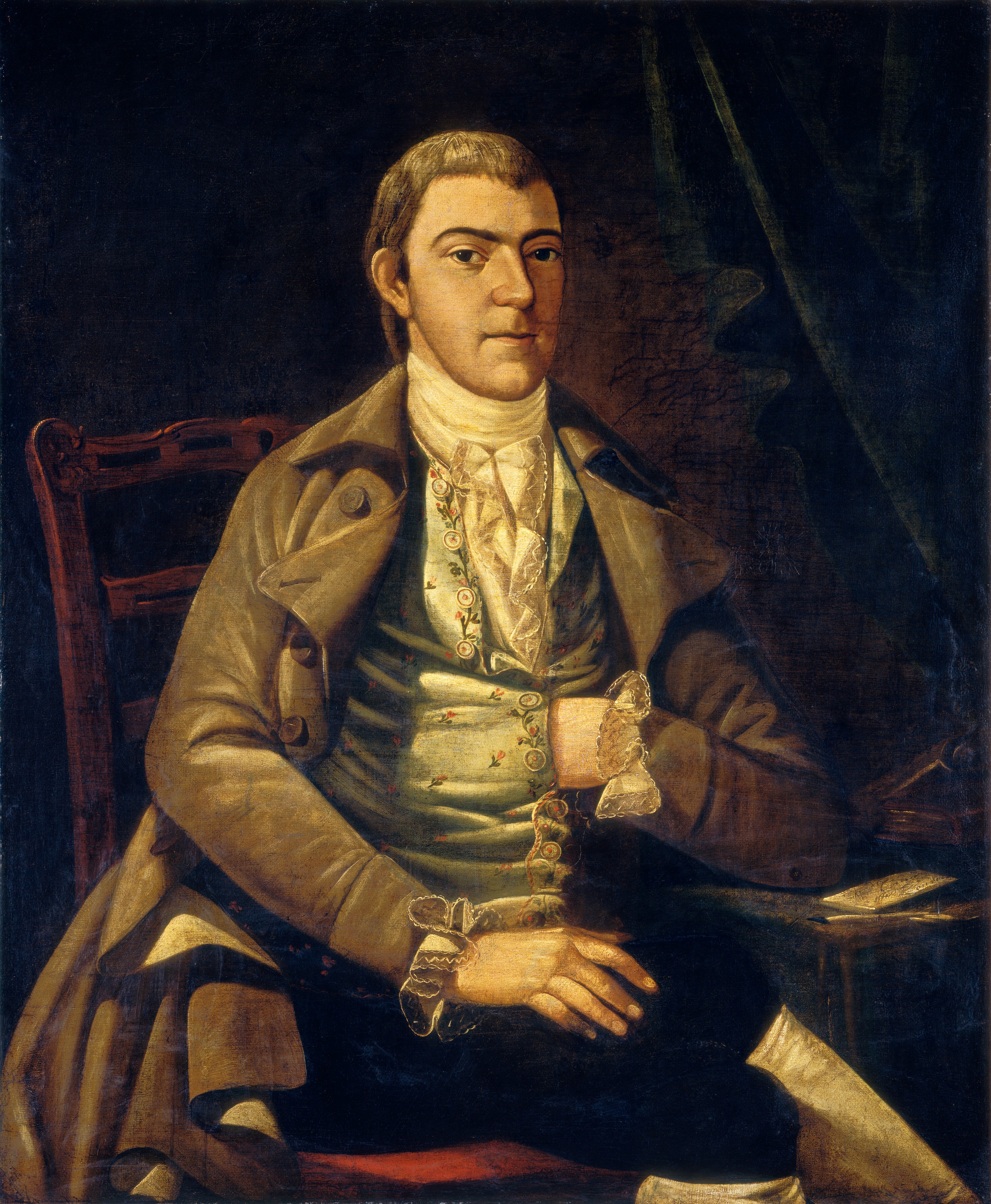 The image shows an upper body portrait of a seated man. He has short brown hair and a firm expression, wearing historical clothing including a brown coat, decorated vest, floral embroidery, and a white cravat. The man has lace cuffs on his sleeves, and is seated on a wooden chair with intricate carvings. His left arm is on his lap, and his right hand is inside his vest. The background is dark with a draped curtain, and a small table with a book or document is to the right of the man.