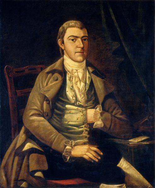 The image shows an upper body portrait of a seated man. He has short brown hair and a firm expression, wearing historical clothing including a brown coat, decorated vest, floral embroidery, and a white cravat. The man has lace cuffs on his sleeves, and is seated on a wooden chair with intricate carvings. His left arm is on his lap, and his right hand is inside his vest. The background is dark with a draped curtain, and a small table with a book or document is to the right of the man.