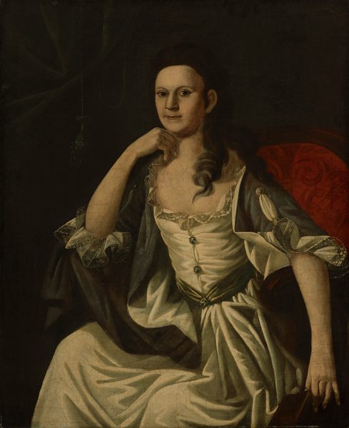 The image depicts a woman seated in a red upholstered chair. She has dark brown, curly hair falling over her left shoulder and is wearing a white dress with a green ribbon tied around the waist. A dark shawl with lace-trimmed sleeves is draped over her shoulders. The woman has a composed expression with softly smiling lips and large, slightly downturned eyes.