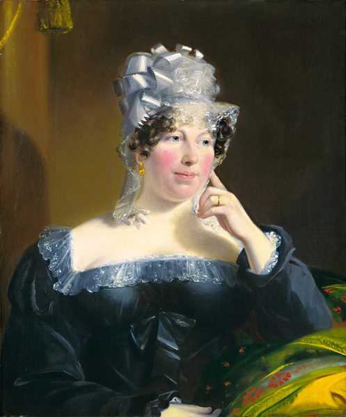 The image shows a woman seated from the waist up, angled slightly to the left. She has fair skin, curly dark brown hair, and is wearing a white bonnet with satin bows. She wears a dark, off-the-shoulder dress with lace trim and a bow, gold earrings, and a ring on her right index finger. On her lap is a colorful fabric with green, yellow, and red hues. The background is a plain, muted dark brown.