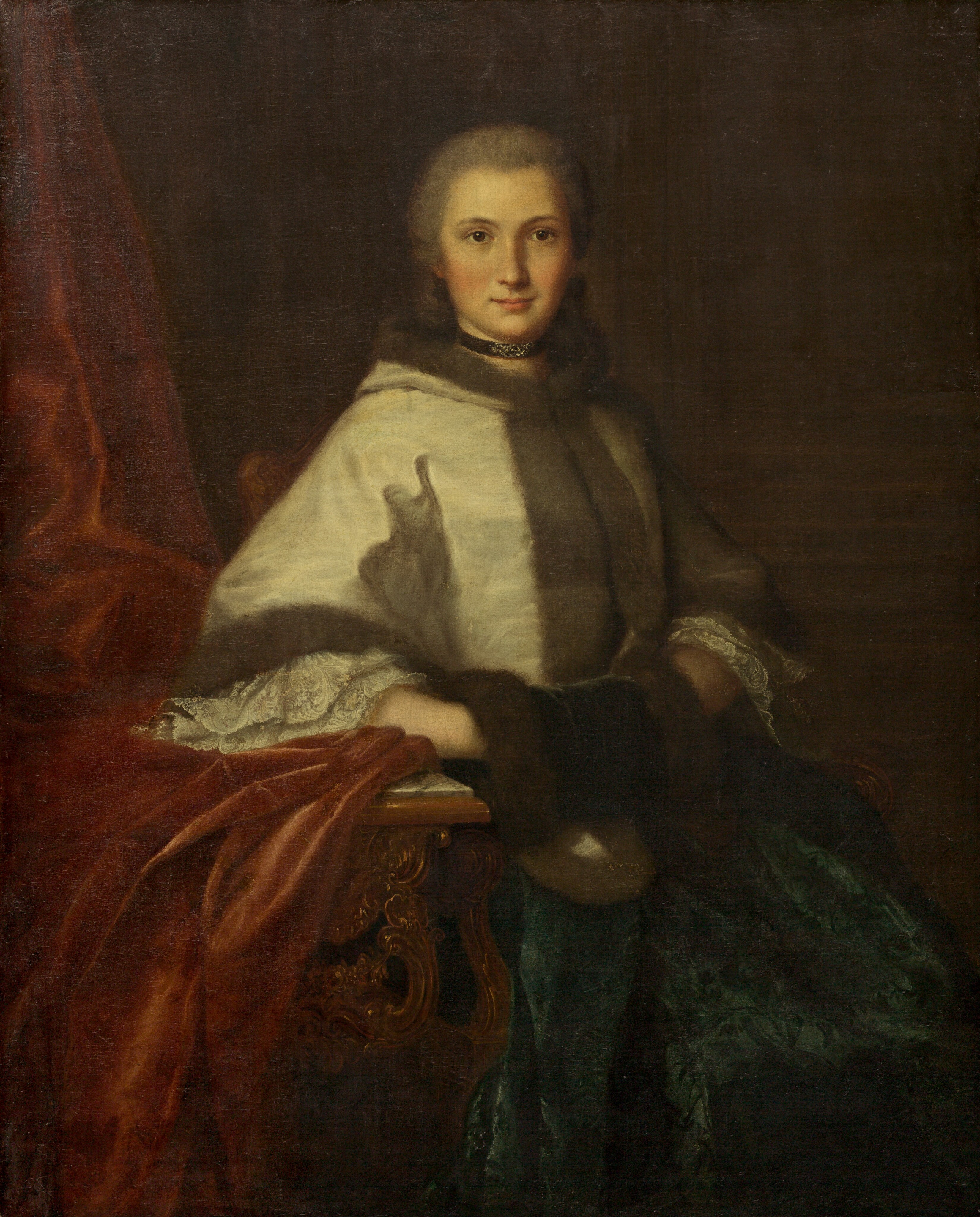 The image displays a woman depicted from the waist up. She is seated, with her body angled slightly to the left, while her head is turned towards the viewer. Her facial features are soft with a serene expression and her hair is pinned back smoothly. She is wearing a luxurious fur-trimmed shawl over a dress with ornate lace cuffs visible at the wrists. The dress features a greenish-blue patterned fabric. Around her neck, she wears a choker necklace. Her hand rests on a wooden table adorned with intricate carvings, and a rich red drapery is elegantly draped over the table's edge. The background is dark and fades into shadow, focusing attention on the subject and her attire.