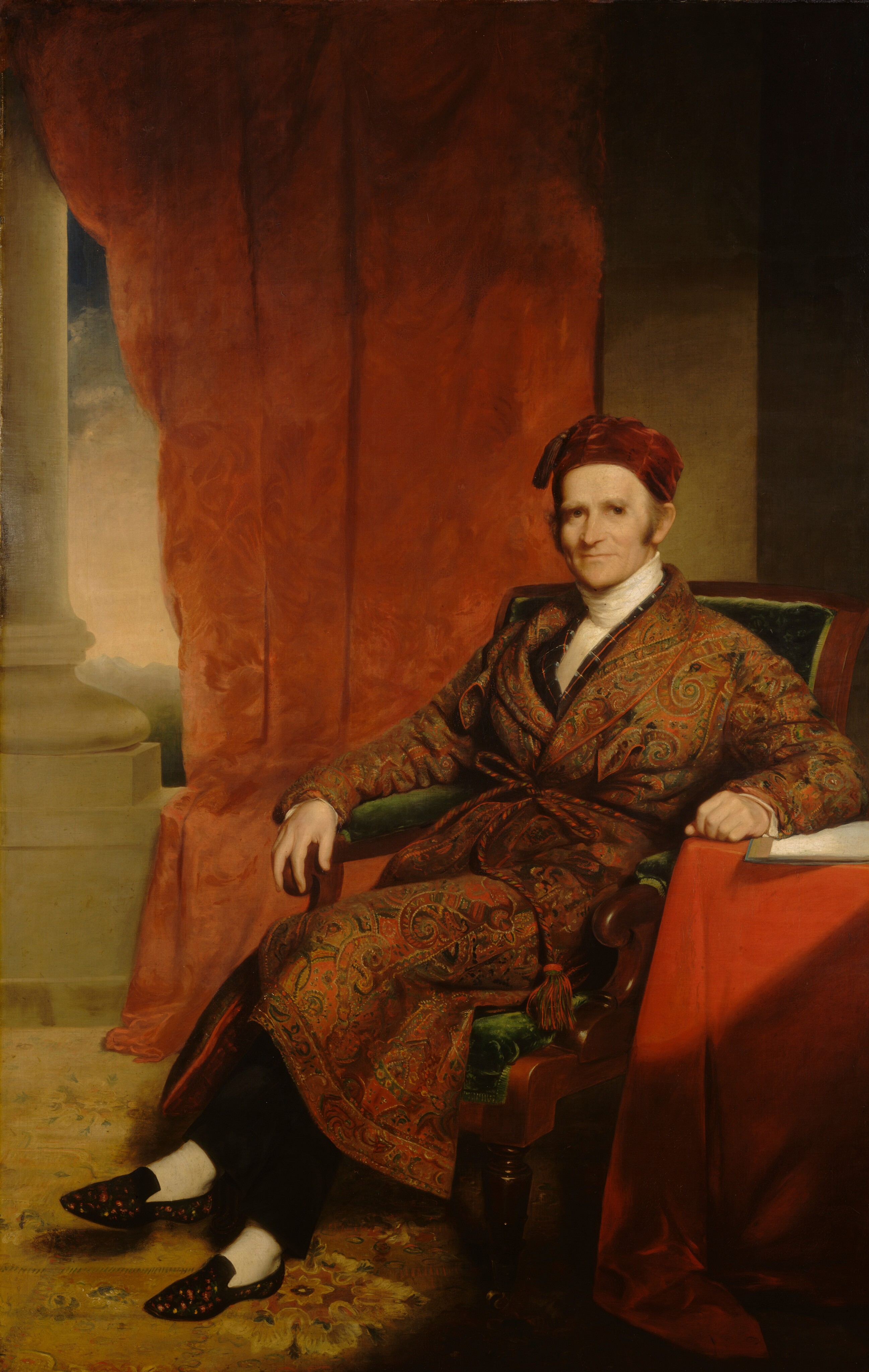 The image shows a man seated on a chair with one arm on the chair's armrest and the other on a table covered with a red cloth. He has fair skin, sideburns, and a smile, and wears a red cap, a patterned robe with a paisley design, dark trousers, white socks, and black slippers with floral embroidery. The background includes red drapery, a column, and a hint of a pale sky.