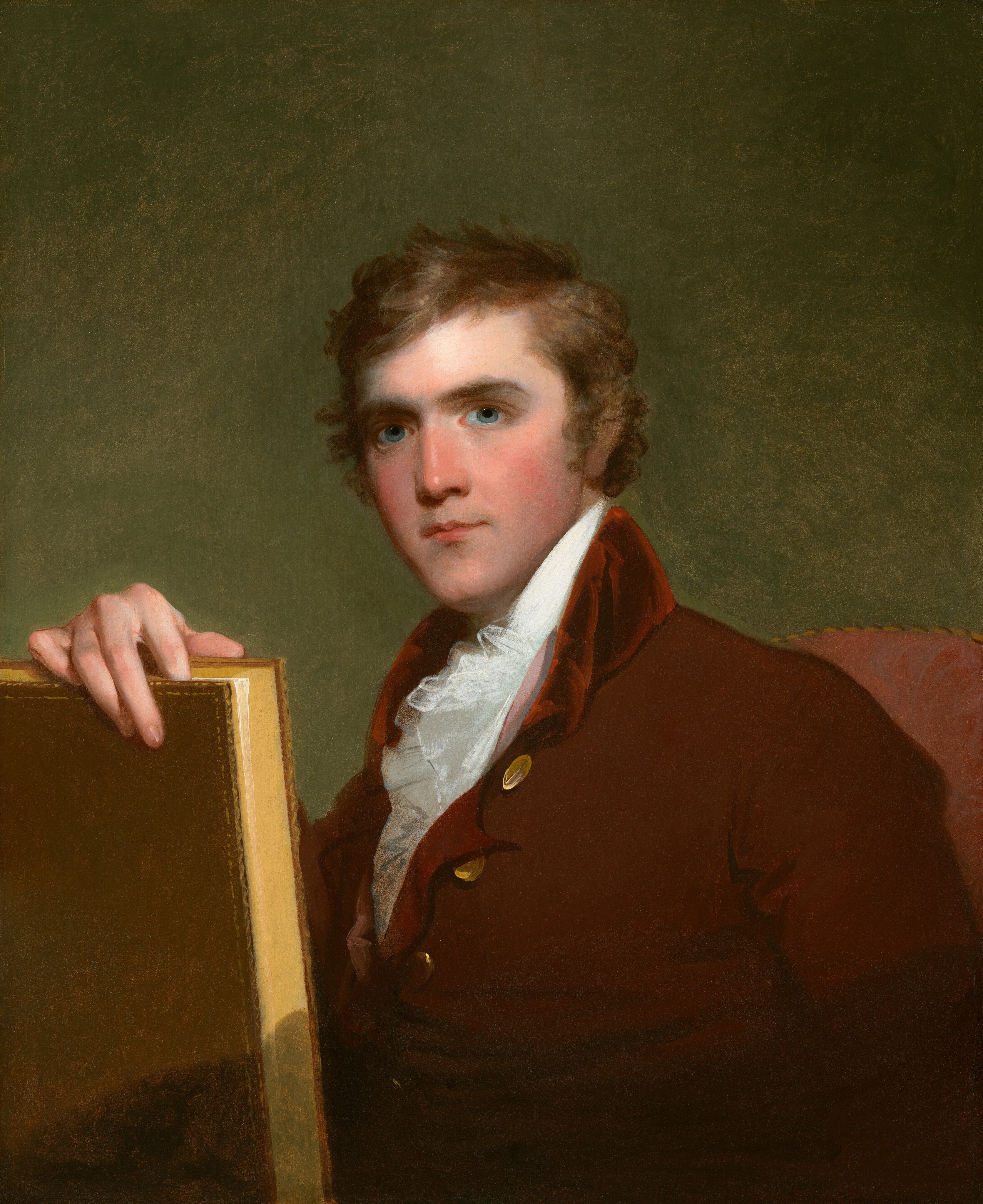 The man in the image is seated and wearing a dark brown coat with brass buttons over a white shirt and ruffled cravat. He has light brown curly hair and is holding a book with a golden cover in his left hand. The background is plain green and the chair he is sitting on is upholstered in red fabric.