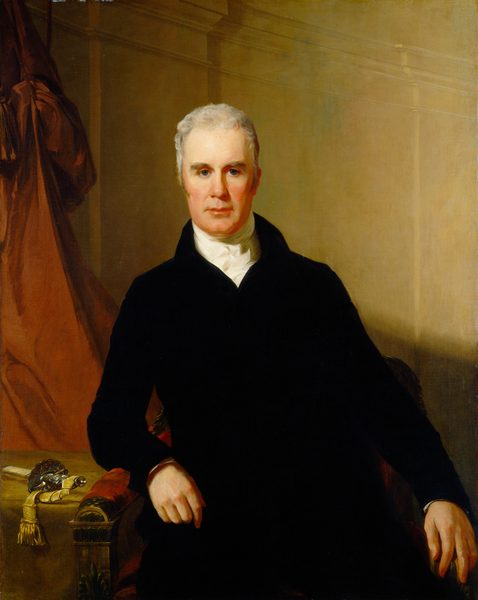 The image shows an upper-body portrait of a man seated in a formal position. The man is depicted with a slight turn of his head, directly facing the viewer with a composed expression. He has short gray hair that is neatly styled. His facial features include a high forehead and firm jawline. He is dressed in a dark formal jacket with a high-collared white shirt visible at the neckline. His left hand rests on the arm of the chair, which is ornately carved with a scroll design. A fabric with an intricate pattern is draped over the back of the chair. There is no visible jewelry. In the background, there is a muted backdrop with a warm hue. On the left side of the image, a flowing drapery in a rich reddish tone adds depth to the setting.