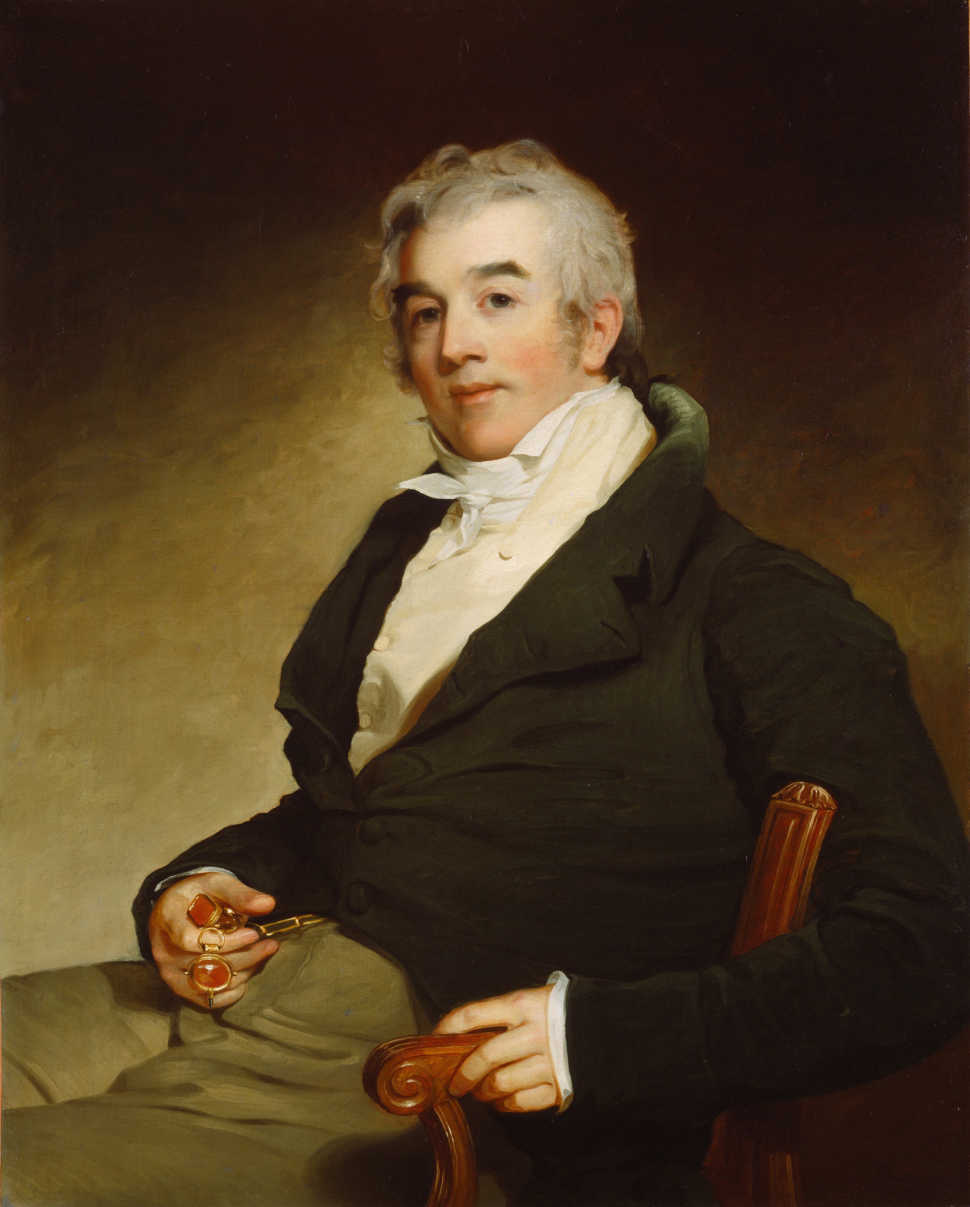 The image depicts a man seated from the waist up. He is slightly turned to the side, with his face towards the viewer. He has a fair complexion, short curly light gray hair, and a composed expression. The man is wearing a dark coat over a white shirt with a cravat. He is holding a pair of spectacles and wearing two large gold rings with polished stones on his fingers. The background is a plain, softly shaded gradient.