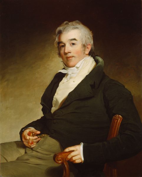 The image depicts a man seated from the waist up. He is slightly turned to the side, with his face towards the viewer. He has a fair complexion, short curly light gray hair, and a composed expression. The man is wearing a dark coat over a white shirt with a cravat. He is holding a pair of spectacles and wearing two large gold rings with polished stones on his fingers. The background is a plain, softly shaded gradient.
