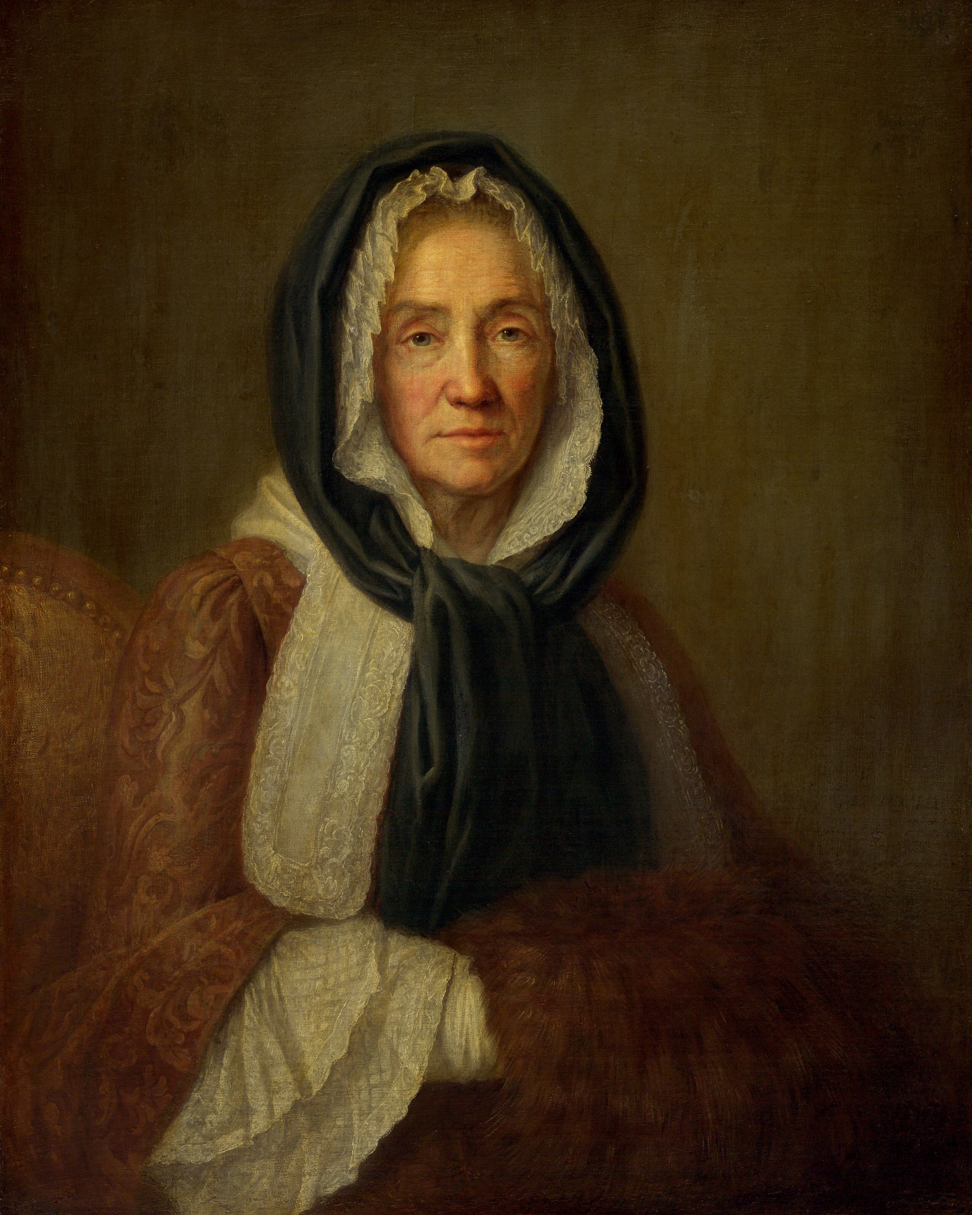 This painting shows a seated elderly woman from the waist up. She faces us with a neutral expression. She has a fair complexion with visible wrinkles, rosy cheeks, and pink lips. Her hair is mostly covered by a white lace cap and a black headscarf. She wears a patterned reddish-brown garment with lace around the collar and sleeves, and her hands rest on a brown fur muff. The background is a dark gray-brown color with visible brushstrokes.
