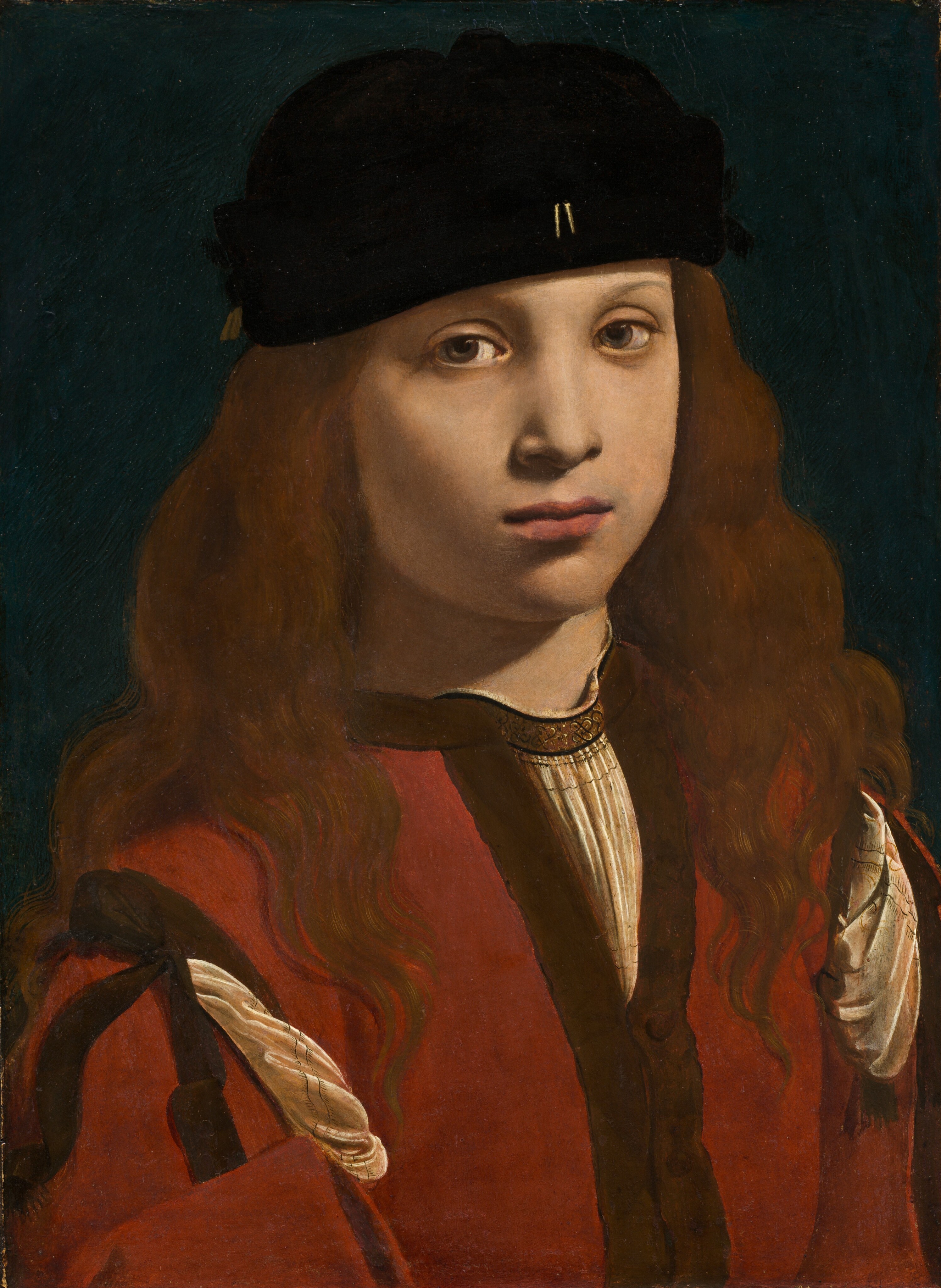 The head, shoulders, and chest of a light-skinned boy or young man wearing a black cap and red garment nearly fill this vertical portrait painting. His body is angled slightly to our right, and he looks at us from the corners of his hazel eyes. He has smooth skin, rounded cheeks, faint, thin eyebrows, and a small nose. His full, dark pink lips are closed. He wears a brimless, soft black cap, slightly askew, over long chestnut-brown hair that covers his ears and flows down past his shoulders. Two small, gold, vertical lines accent the front center of his cap near the lower edge. He wears a collarless, crimson-red jacket, with dark brown trim along the edges and at the shoulder. Pleated folds of his ivory-white shirt material puff out from openings at the shoulders and a V at the top of the chest. There is a also brown band around the neck of the white shirt with a pattern of black, knot-like designs. The white shirt also has two rows of delicate black lines where it is pulled through at the shoulders. The boy is lit from our right and above, casting a shadow on his neck and cheek to our left. The background behind him is teal blue.