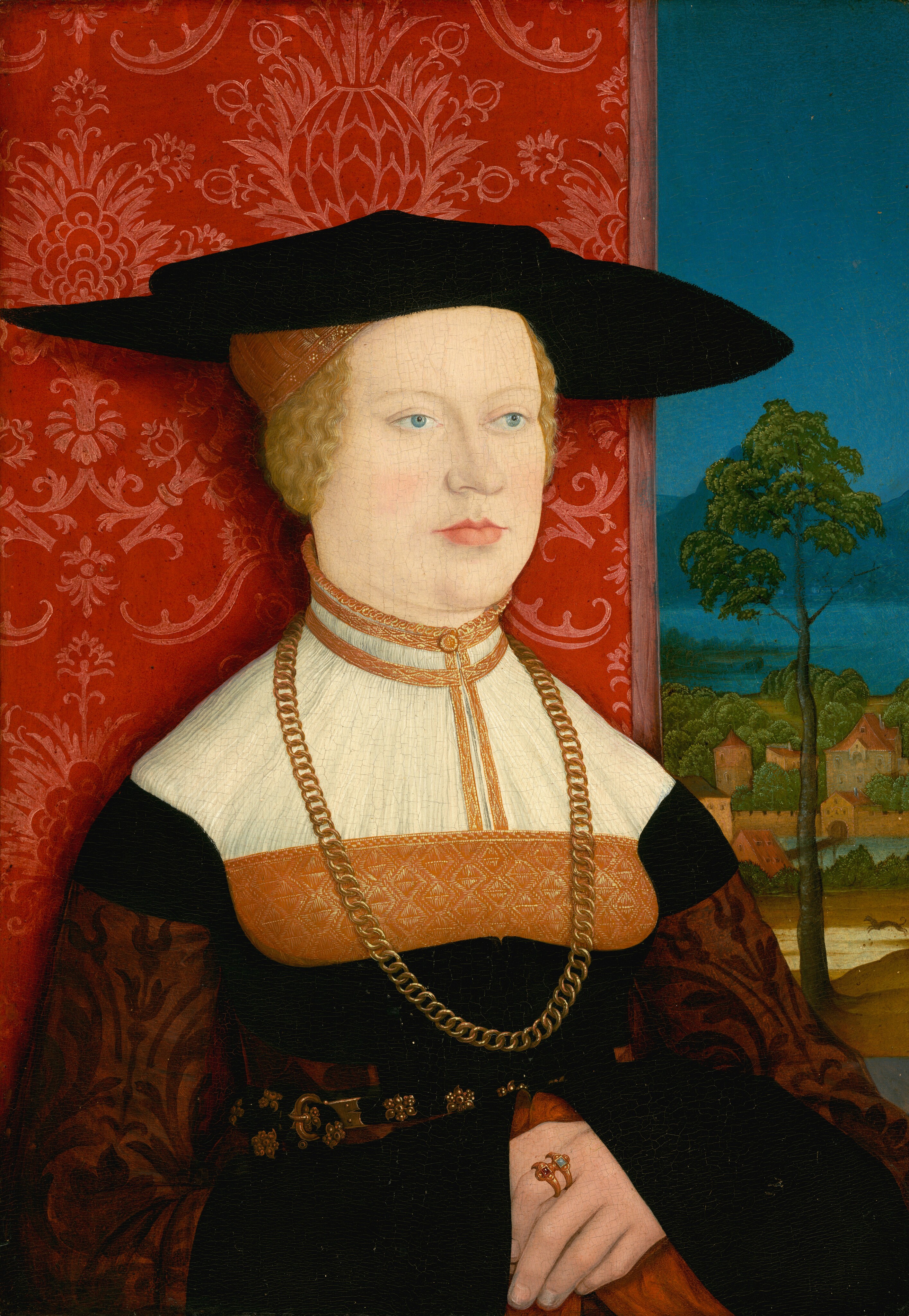 Shown from the waist up, a light-skinned woman sits angled slightly to our right in this vertical portrait painting. The woman has blue eyes, faint brows, and a sloping nose ending in a small bulb. Her upper lip is thin and her lower lip full over a slight double chin. Her blond hair is pulled back under a gold, cap-like headdress and a black, wide-brimmed hat with a shallow crown. She wears a high-necked white, black, and gold dress with intricate detailing, a gold chain necklace that falls to her waist, and a black belt with a gold clasp and medallions. She wears two gold rings set with gems on the pointer finger of her left hand, which is folded across her belly over the other hand. A scarlet-red wallcovering with pink floral designs fills two-thirds of the space behind her. The rightmost third of the background opens onto a landscape with a tall, spindly tree, a leaping animal, presumably a dog, and a bridge across a body of water leading to a town. Across another body of water, perhaps a lake, mountains are marine blue in the distance under a clear, celestial-blue sky.