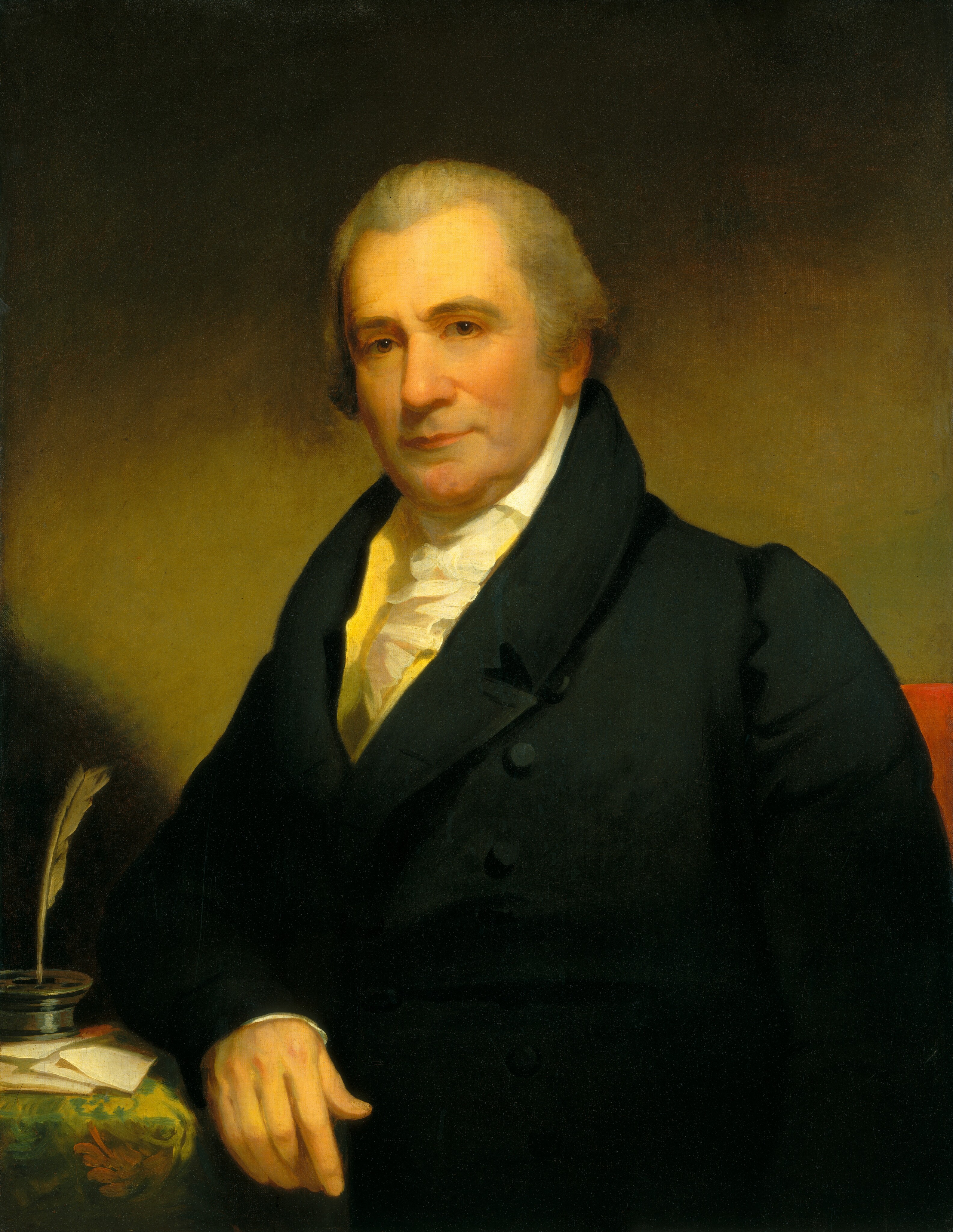 The image shows a man sitting and facing the viewer. He has his right hand on a table. The man's hair is gray and neatly styled. He is wearing a dark coat with a high collar, a white shirt, and a yellow vest peeking through. On the table are a quill, inkwell, and papers. The background is a dark, neutral tone.